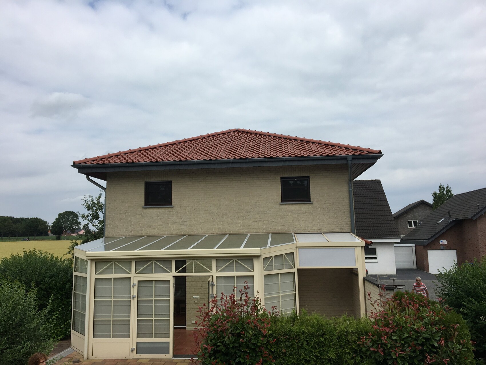 Zweistöckiges beige Ziegelhaus mit rotem Ziegeldach, Glasveranda und Garten.