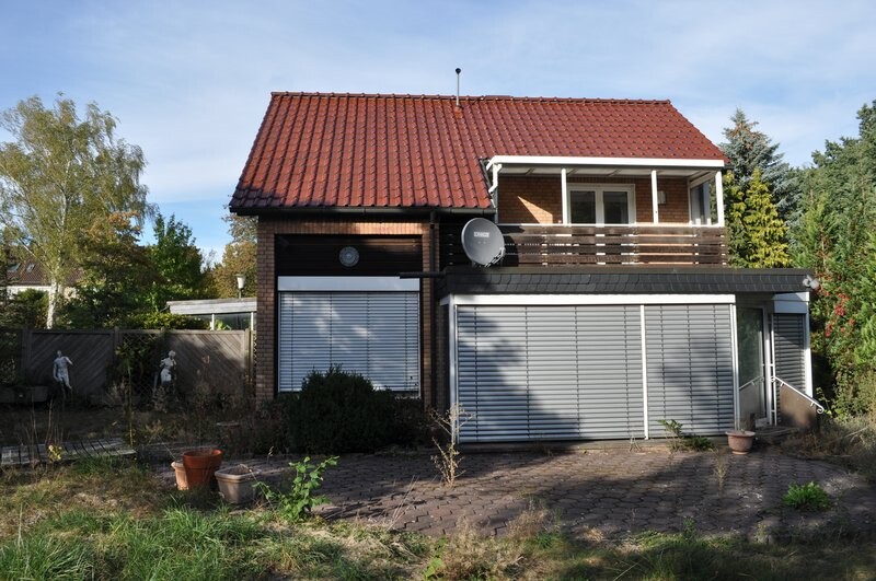 Zweistöckiges Backsteinhaus mit rotem Dach, Rollläden, Balkon und gepflastertem Hof.