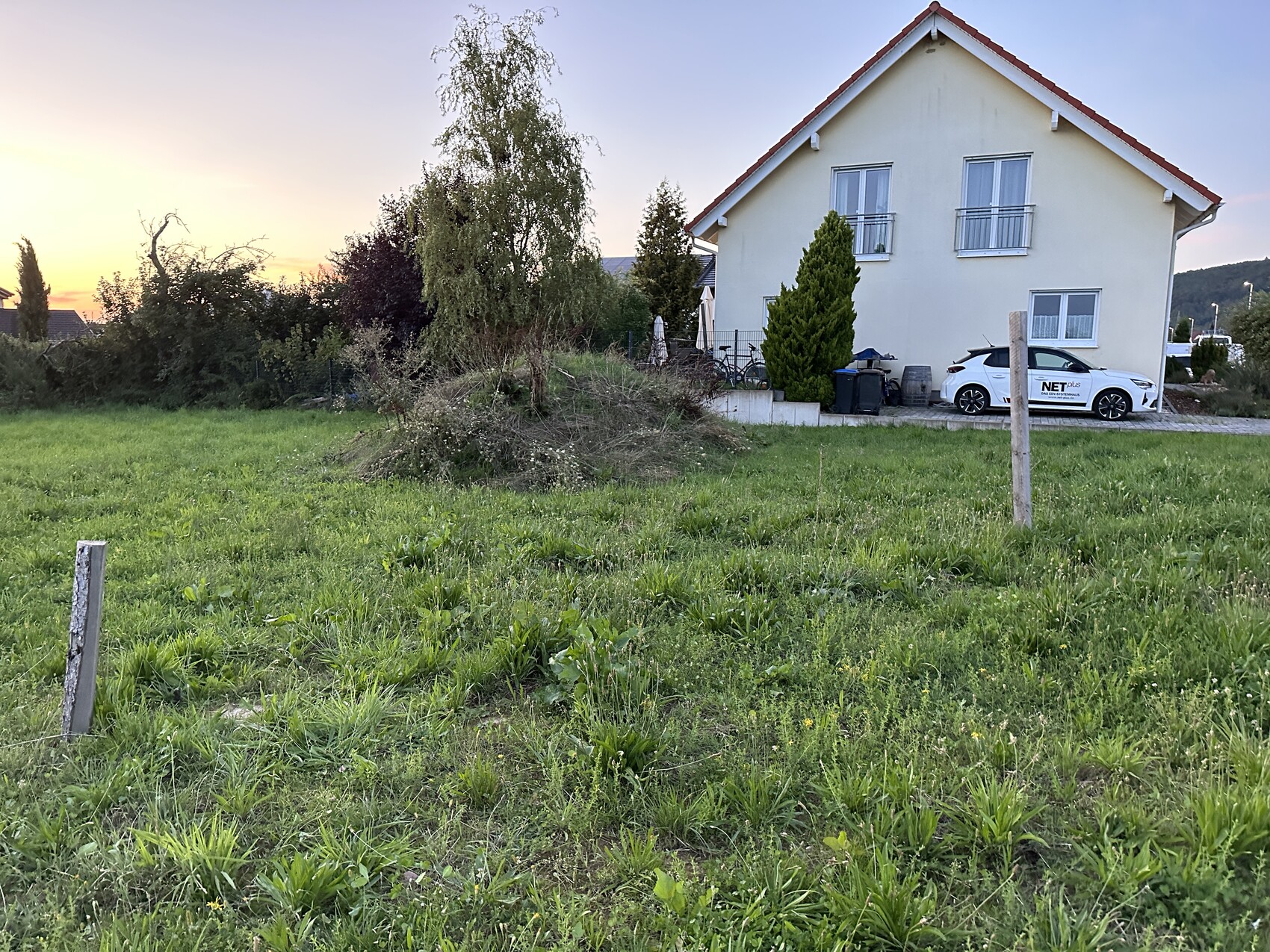 Weißes zweistöckiges Haus mit rotem Dach rechts, davor grüne Wiese und geparktes Auto.