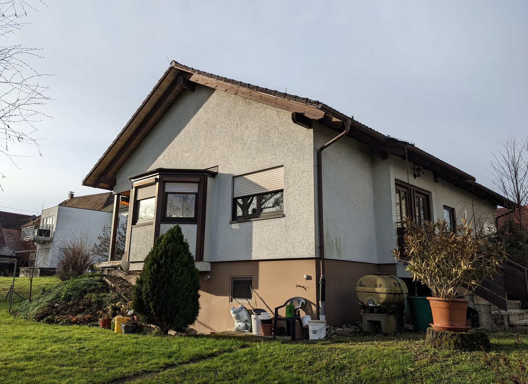 Einfamilienhaus mit grauer Putzfassade, braunem Dach und Gartenpflanzen im Vordergrund.