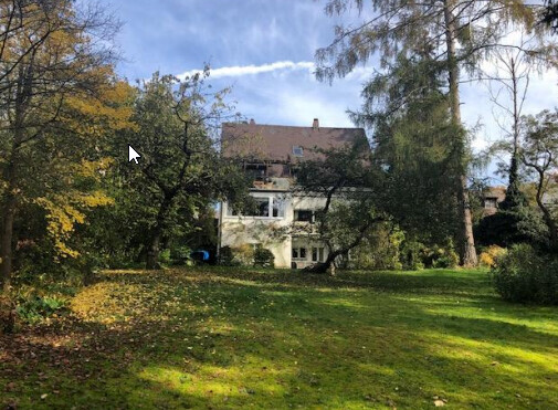 Zweistöckiges Haus im Garten, von Bäumen eingerahmt, Herbstlaub auf dem Rasen, blauer Himmel.