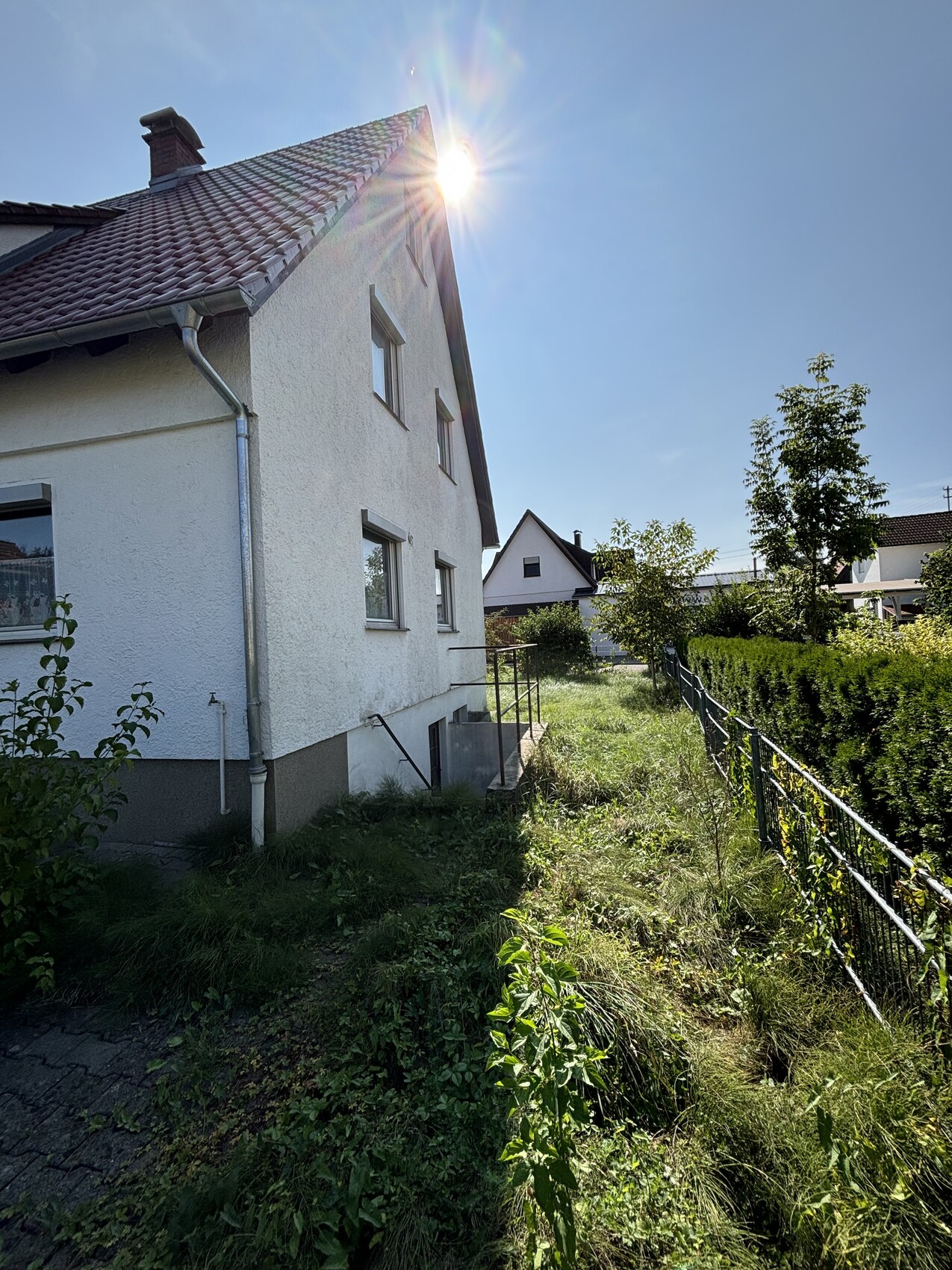 Weiße Hausfassade mit rotem Dach, sonnigem Himmel, Garten und Zaun am Rand