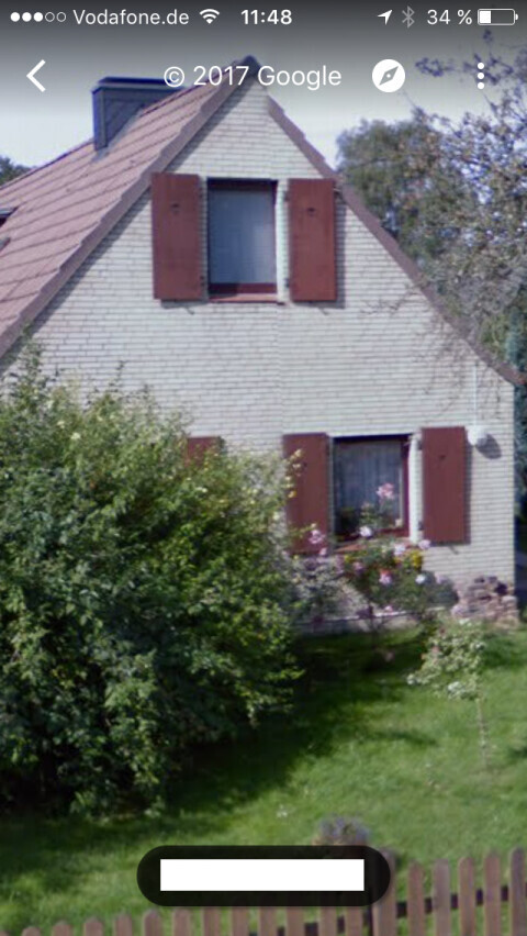 Zweigeschossiges Haus mit braunen Fensterlaeden, Garten und Zaun im Vordergrund
