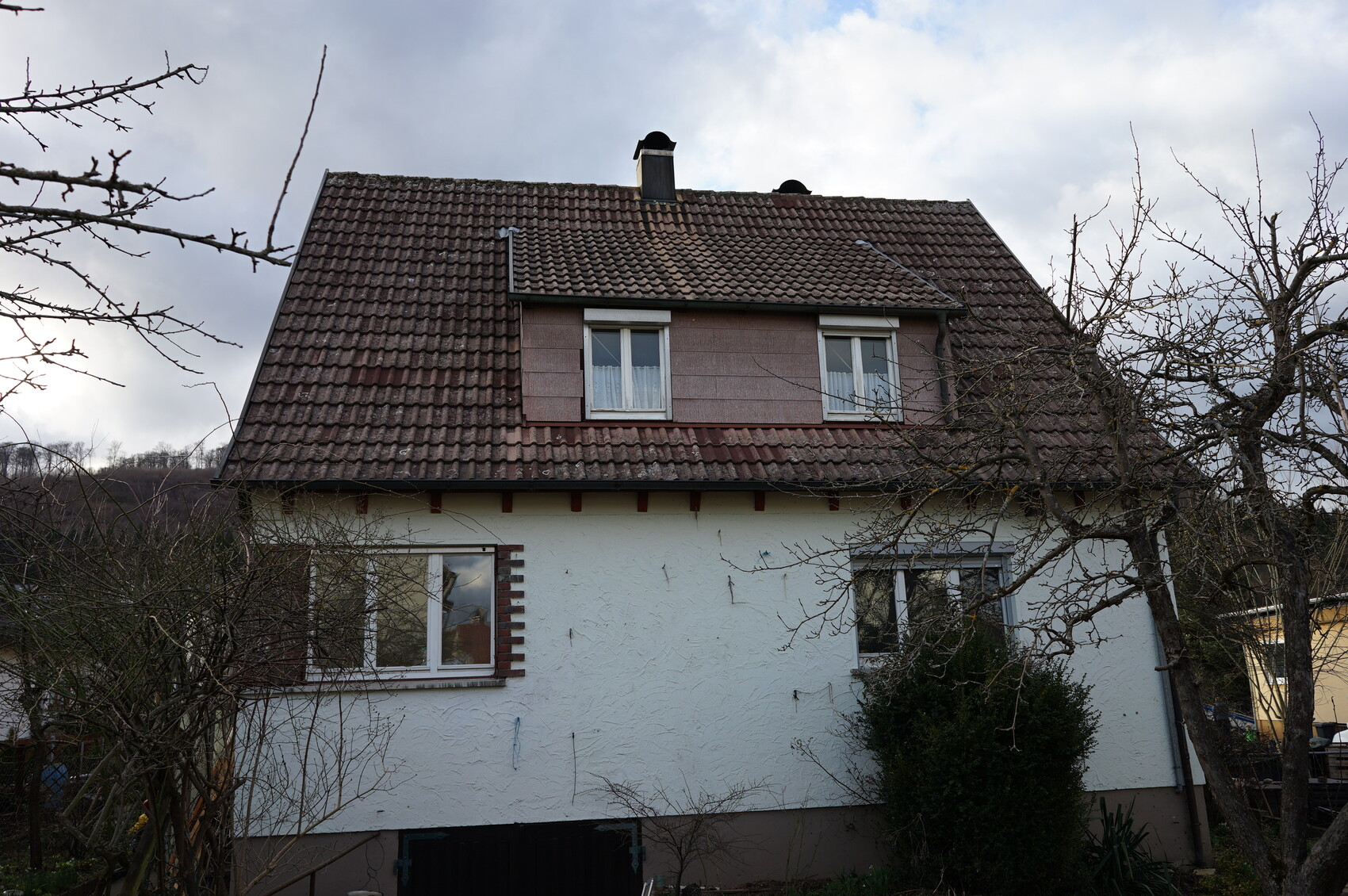 Zweistöckiges Haus mit rotem Ziegeldach und zwei Dachfenstern, von kahlen Bäumen umgeben.