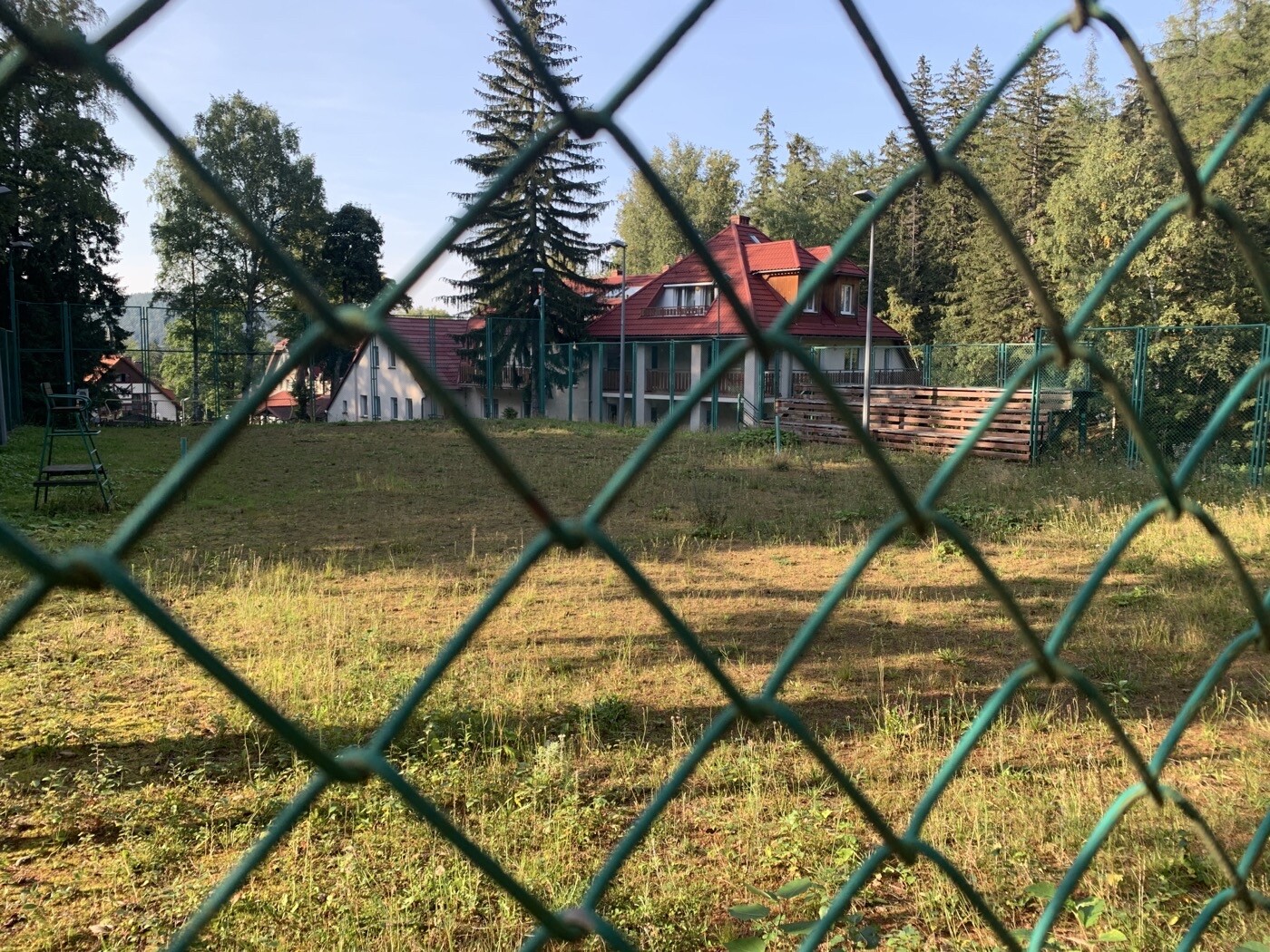 Grüner Kettenzaun im Vordergrund, freies Feld, dahinter Häuser mit roten Dächern.