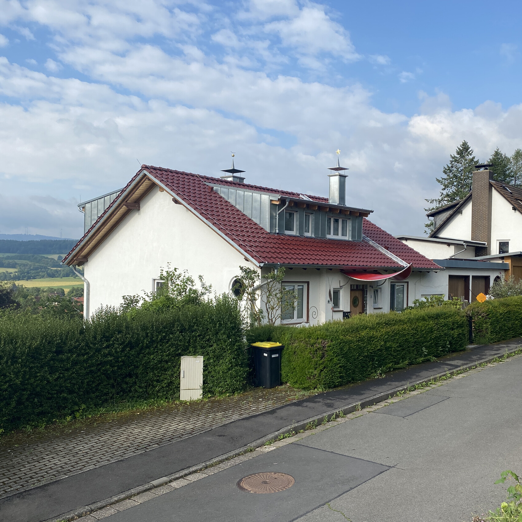 Aussenansicht eines Einfamilienhauses mit weisser Fassade rotem Dach und gruener Hecke.