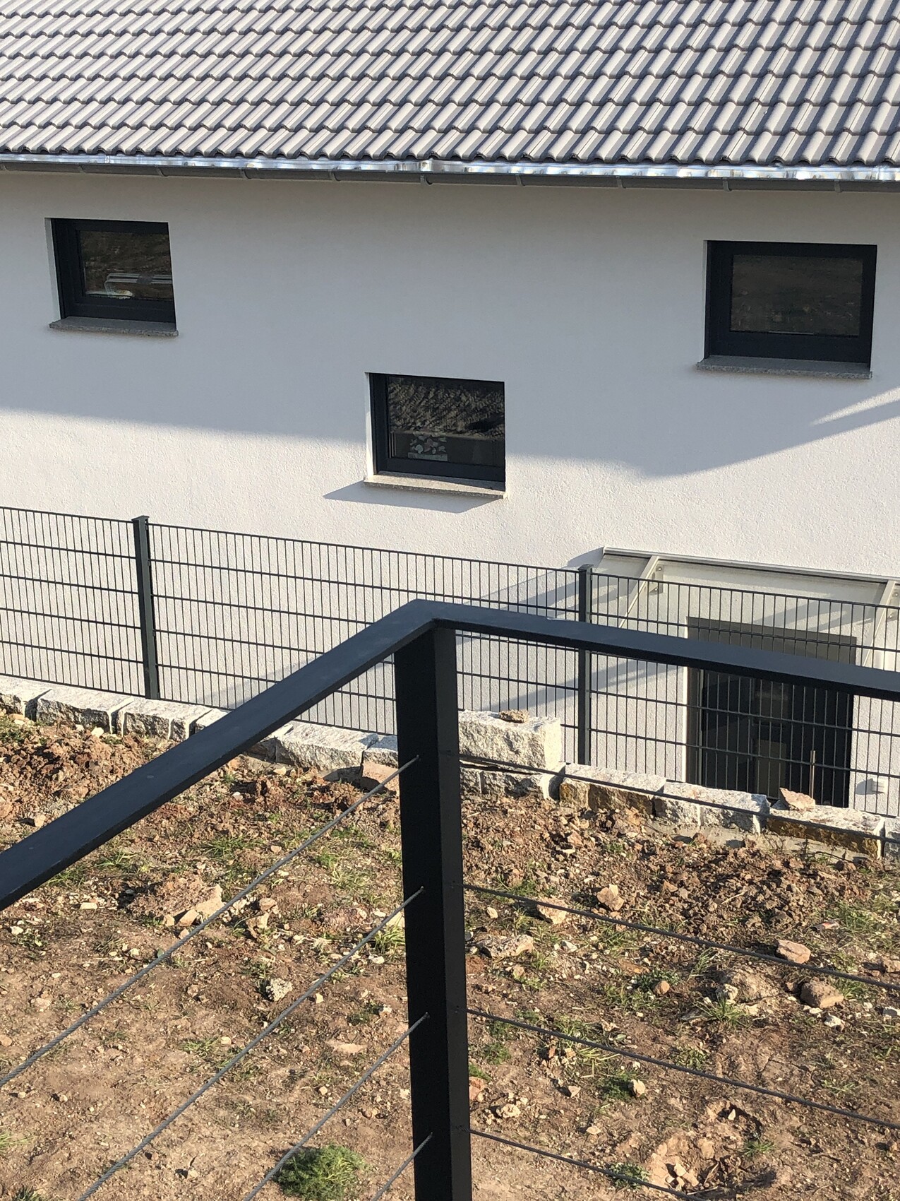 Außenansicht eines weißen Hauses mit drei dunklen Fensterrahmen; schwarzes Geländer im Vordergrund.