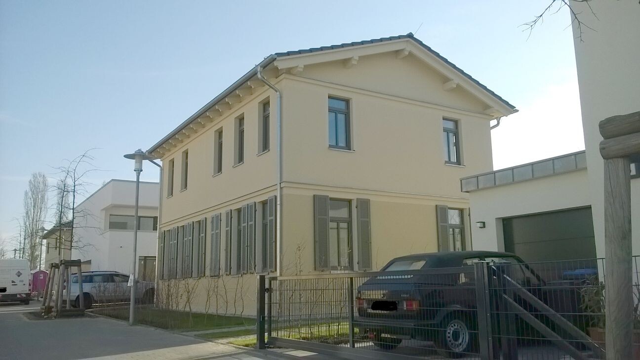 Zweistöckiges, hellbeiges Haus mit Rollläden; vor dem Zaun parkt ein Auto.