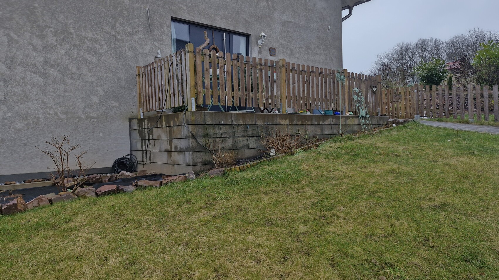 Außenansicht eines Hauses mit Holzlattenzaun, erhöhter Terrasse, Gartenschlauch und Gras.
