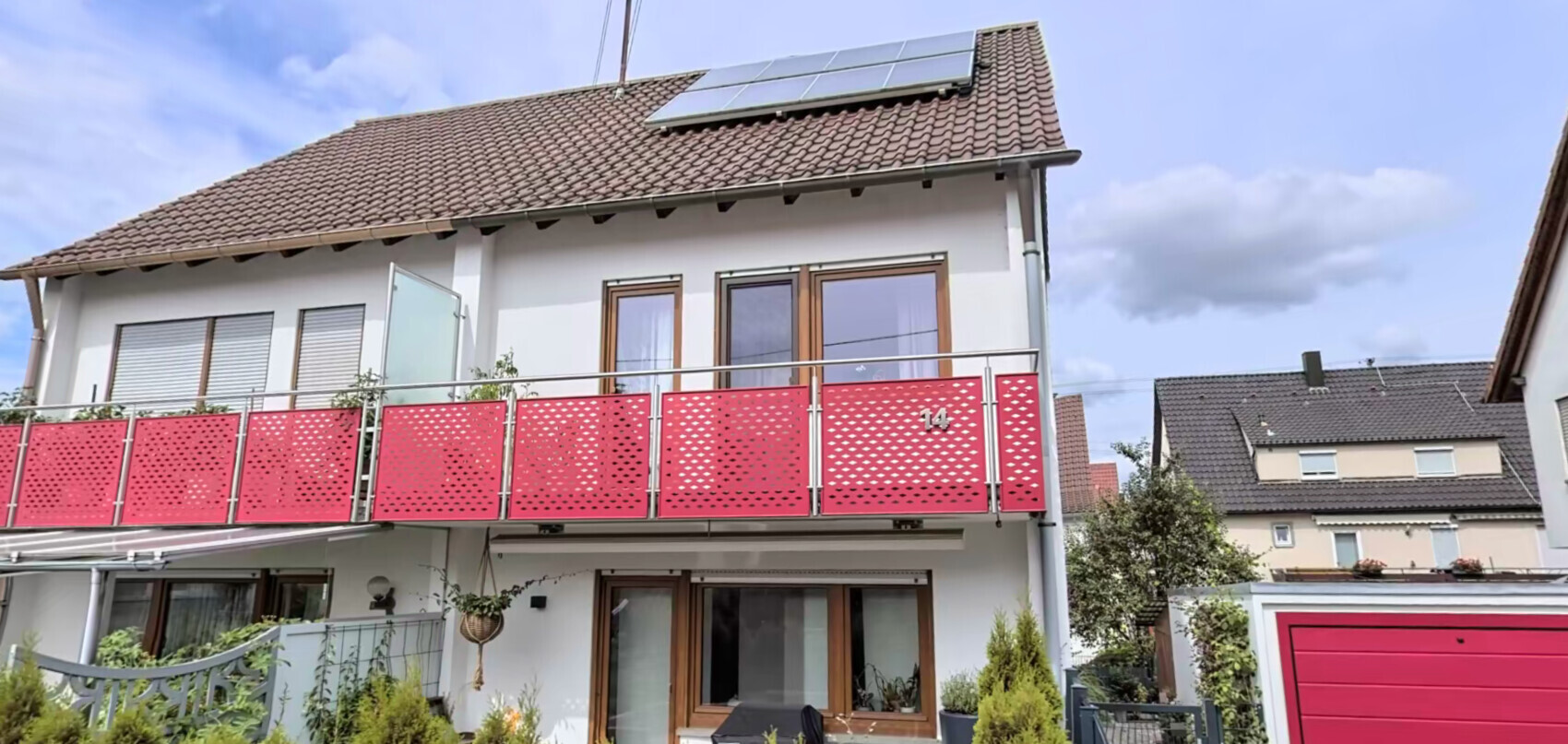 Zweistöckiges Haus mit rotem Balkon aus perforierten Paneelen und Solarpaneelen auf dem Dach