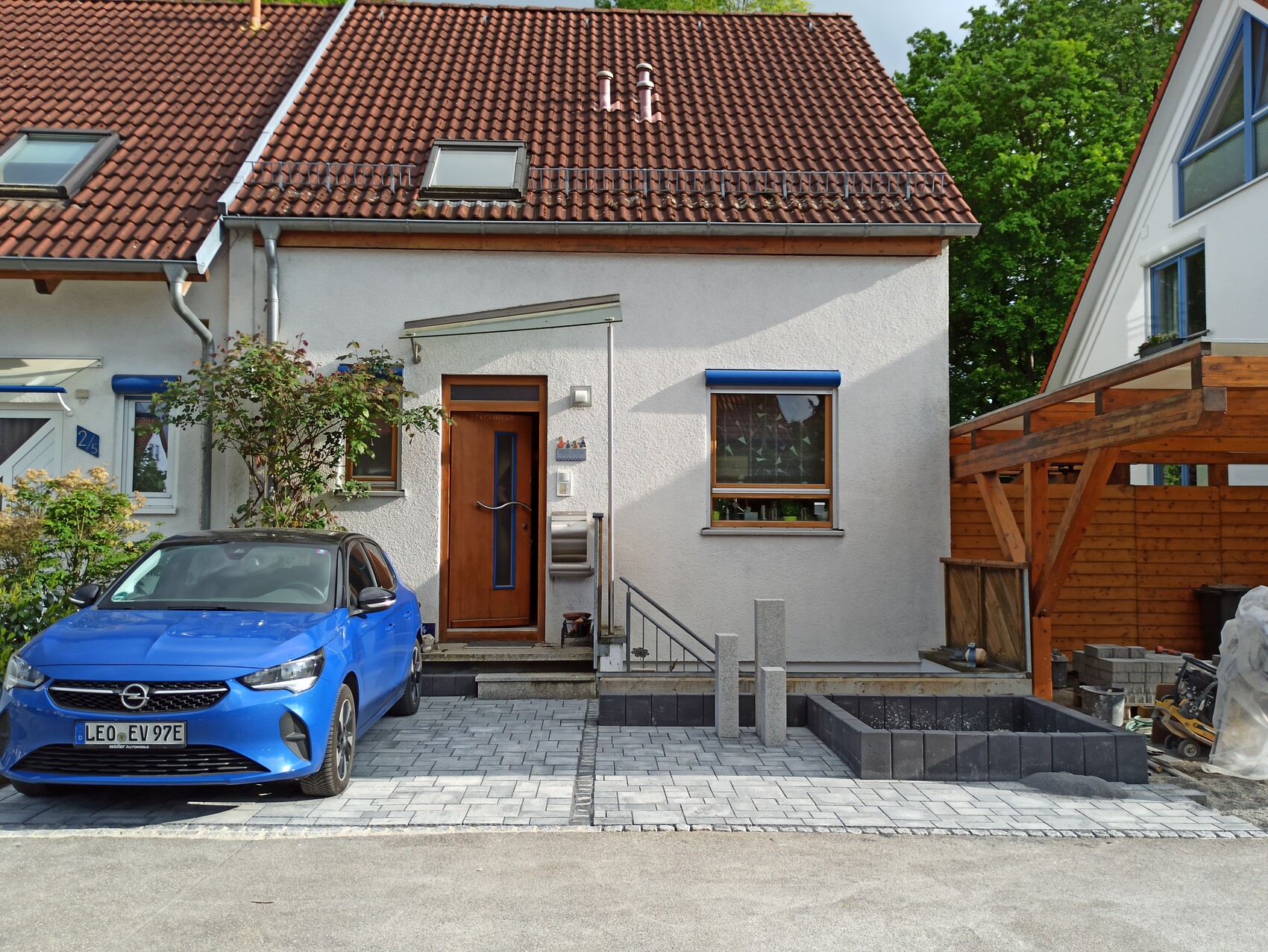 Weiße Hausfront mit rotem Ziegeldach, blaues Auto vor Eingang, Baubereich rechts.