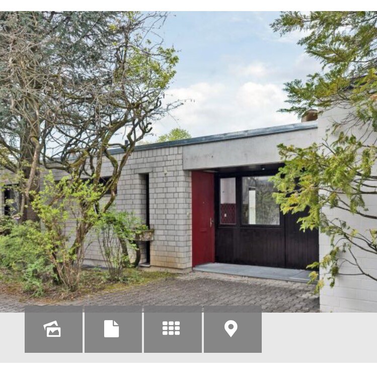 Außenansicht eines modernen Hauses mit grauer Wand, roter Tür und angrenzenden Bäumen.