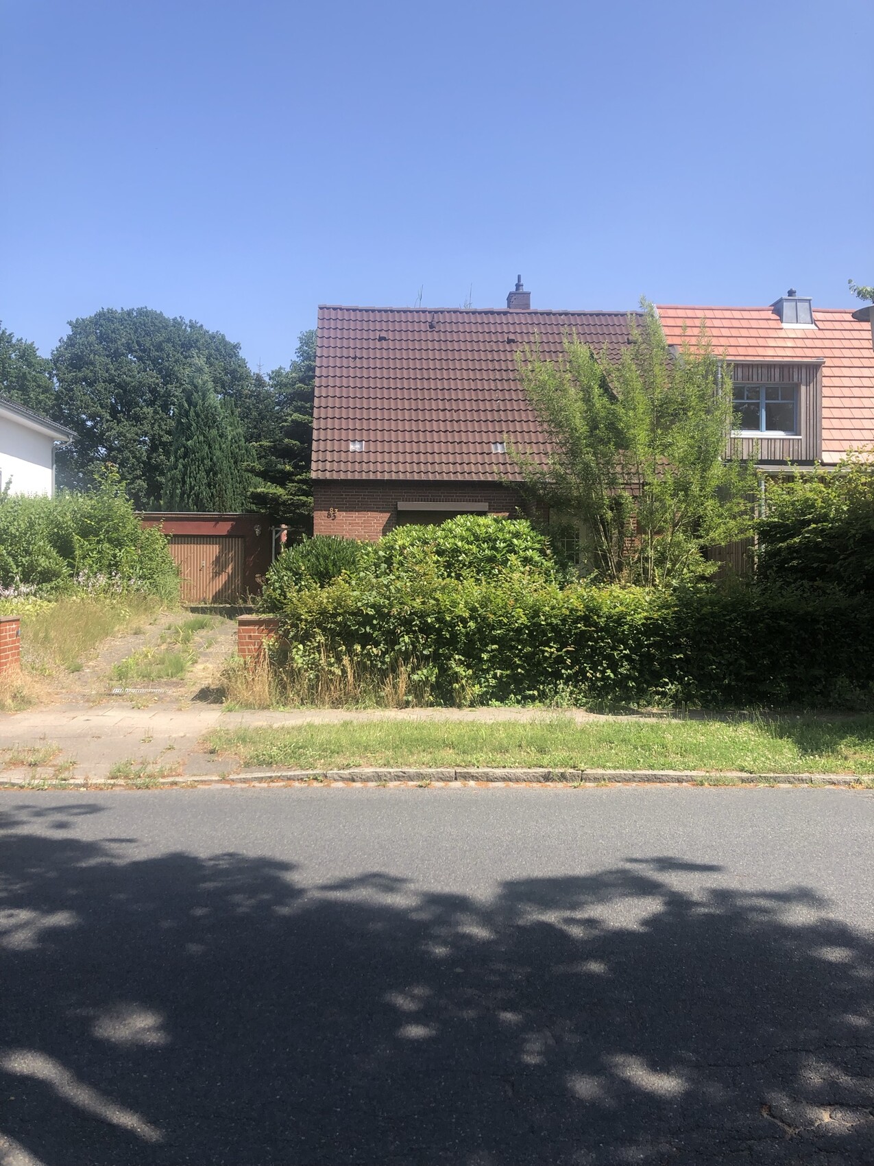 Haus mit rotem Ziegeldach, grüne Hecke im Vorgarten, Straßenzug davor, blauer Himmel