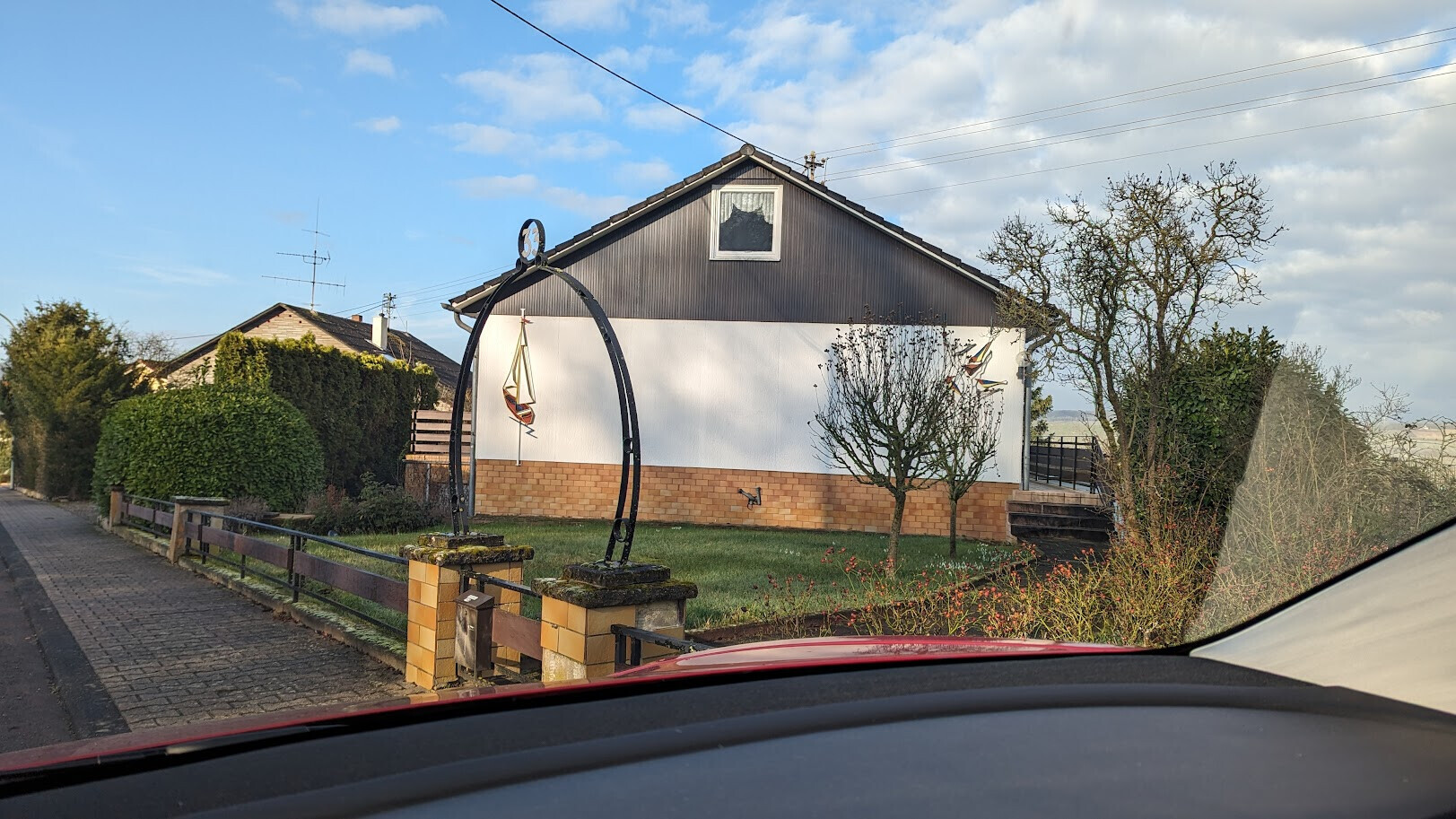 Wohnhaus mit weißer Wand, braunem Sockel, Garten und Metallbogen mit Segelbootmodell im Vorgarten