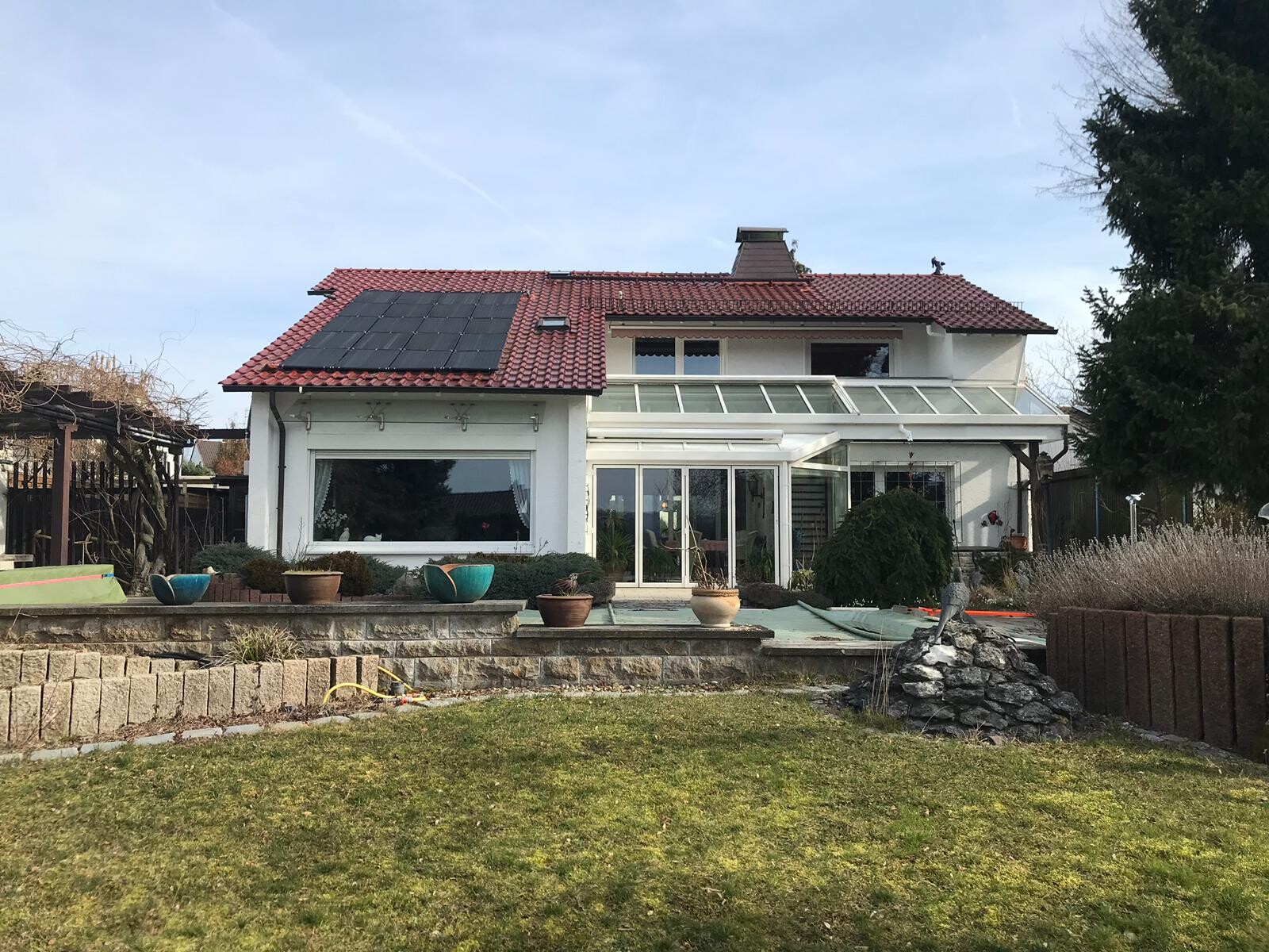 Außenansicht eines Einfamilienhauses mit rotem Dach, Solarpanelen, Glasveranda und Garten.