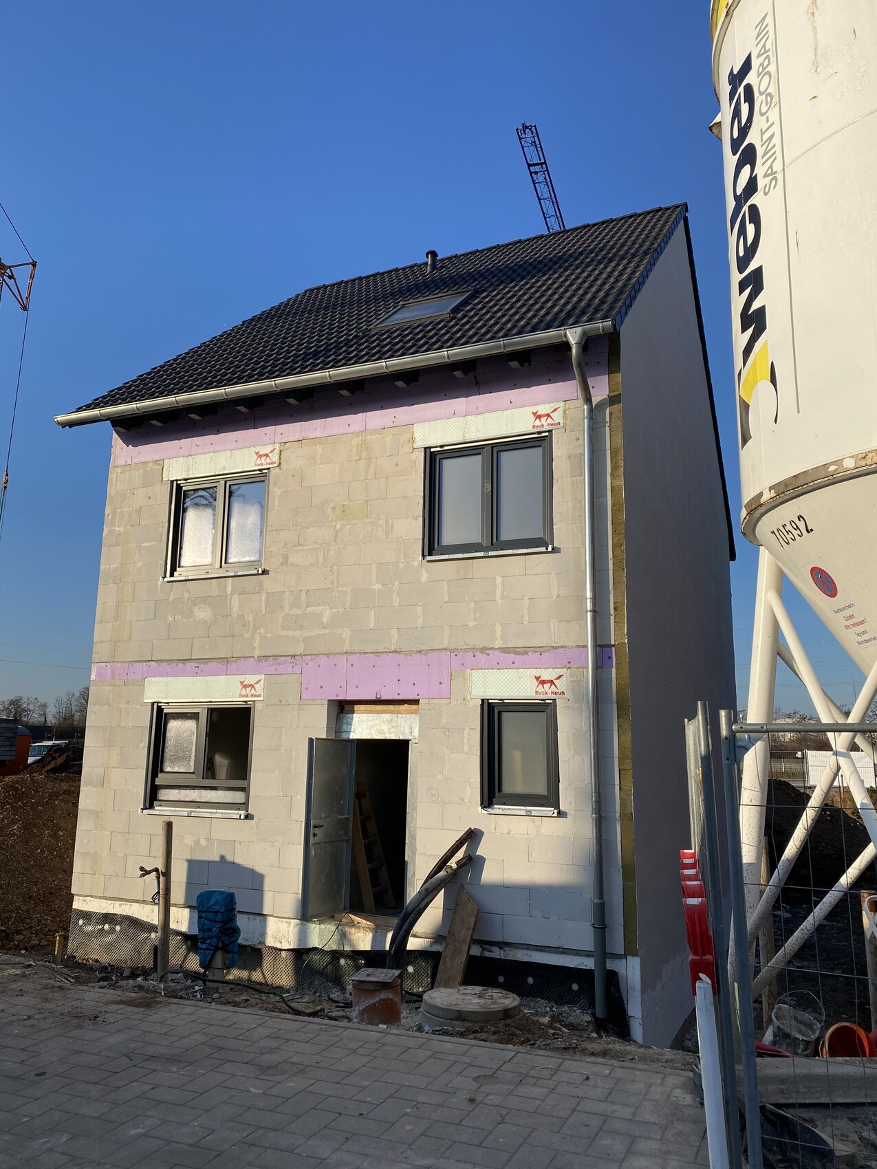 Zweistöckiges Haus im Rohbau aus Betonblöcken mit offener Eingangstür und Baugeräten.