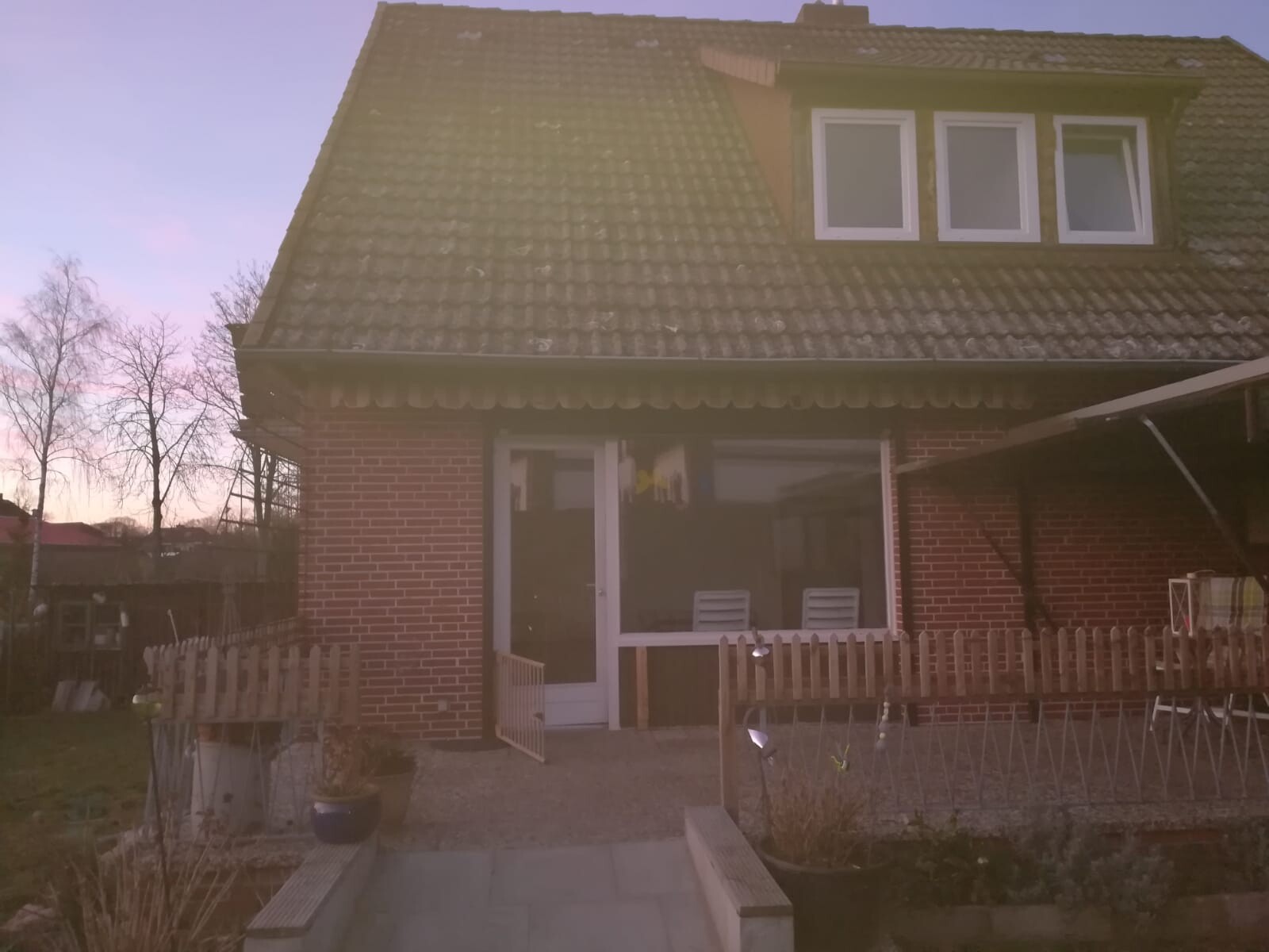 Frontansicht eines roten Backsteinhauses mit Dachgauben, Glasfront und Gartenzaun.