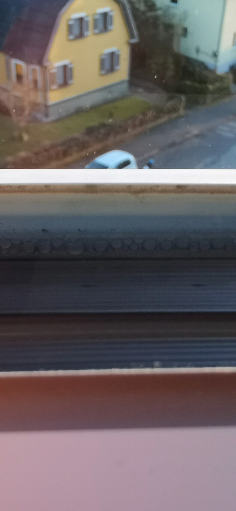 Blick durch schmutziges Fenster auf gelbes Haus mit weißen Fenstern und vorbeifahrendem Auto.