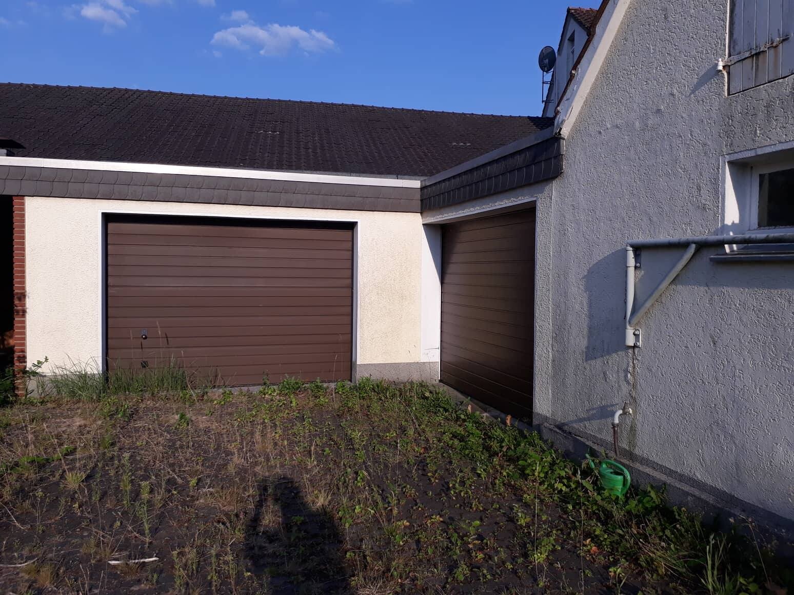 Außenansicht eines Hauses mit zwei braunen Garagen-Toren und ungepflegtem Vorgarten