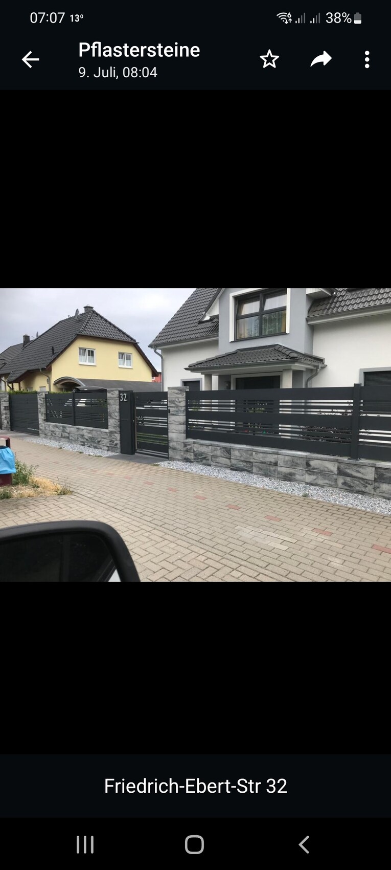 Moderne Einfamilienhausfront mit grauem Dach, schwarzem Lamellenzaun und Steinmauer am Gehweg Nr. 32.