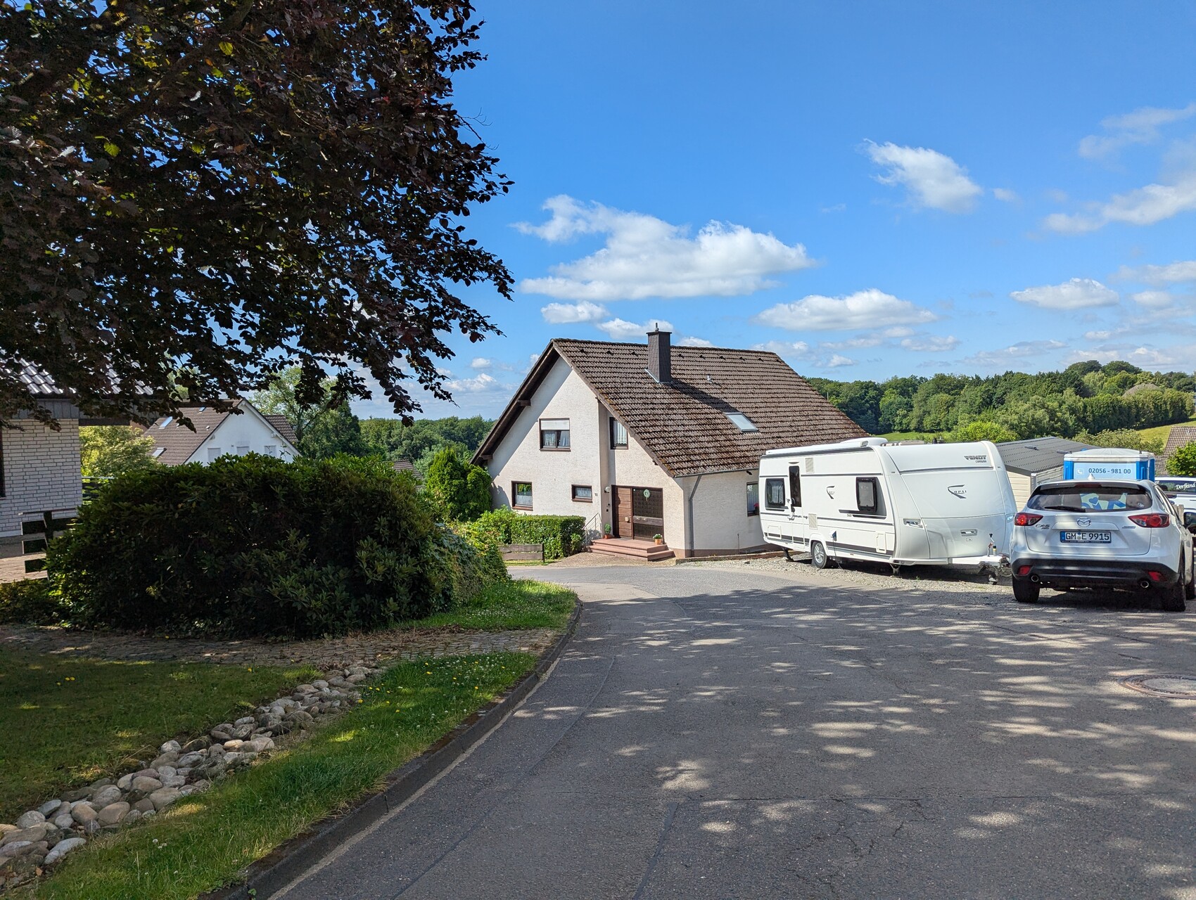 Weißes Einfamilienhaus mit steilem Dach, davor Wohnwagen und Auto; ländliche Umgebung.
