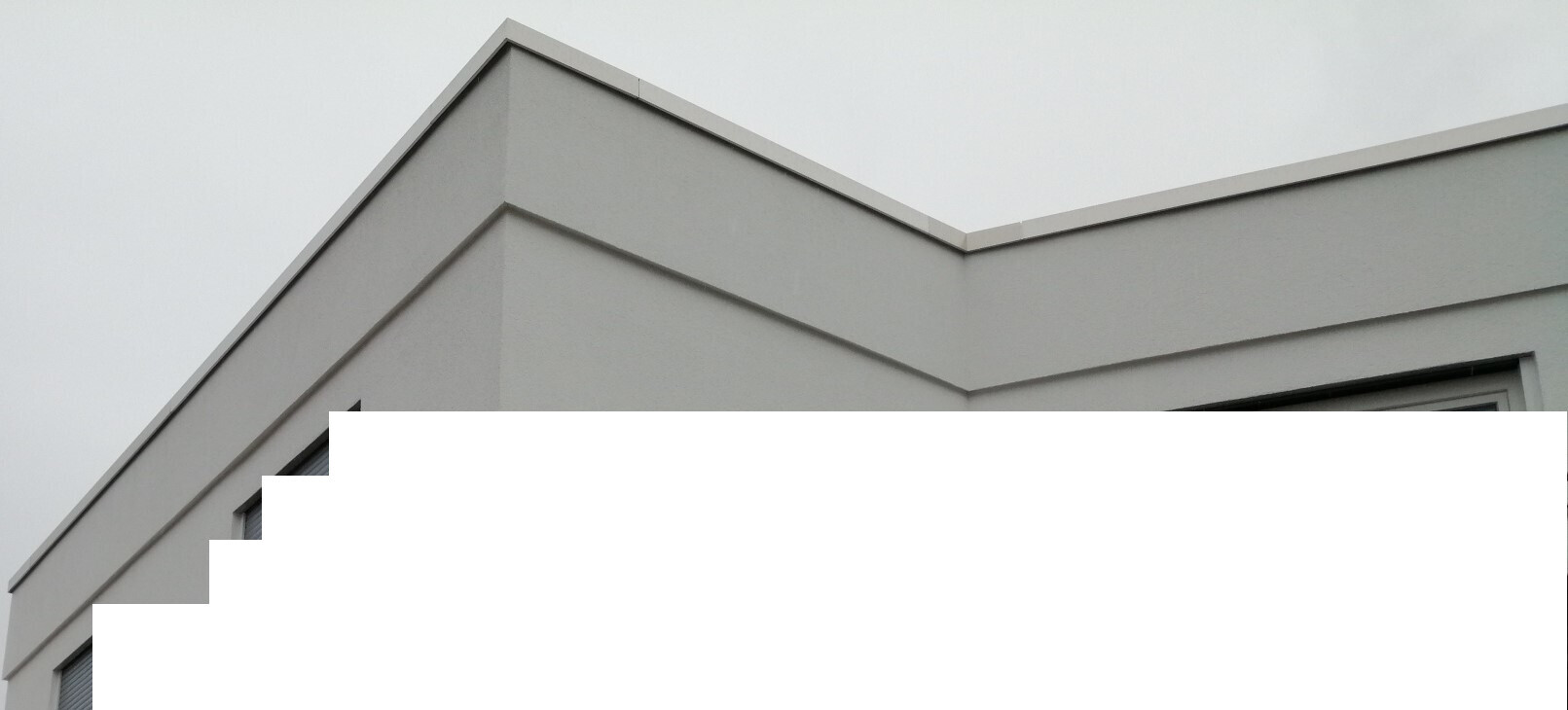 Moderne graue Gebäudeecke mit gestaffelten Fassadenkanten und scharfem Dachrand vor wolkigem Himmel.