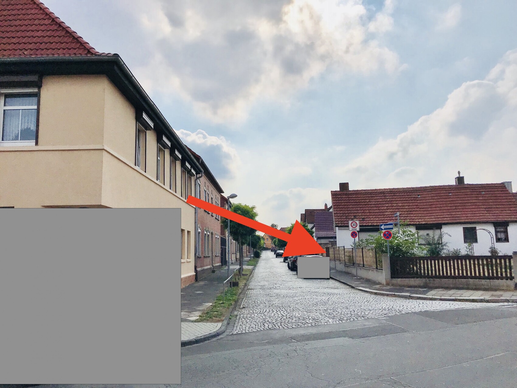 Straßenansicht mit Reihenhäusern links, Einfamilienhäuser rechts, gepflasterte Straße und Wolken.