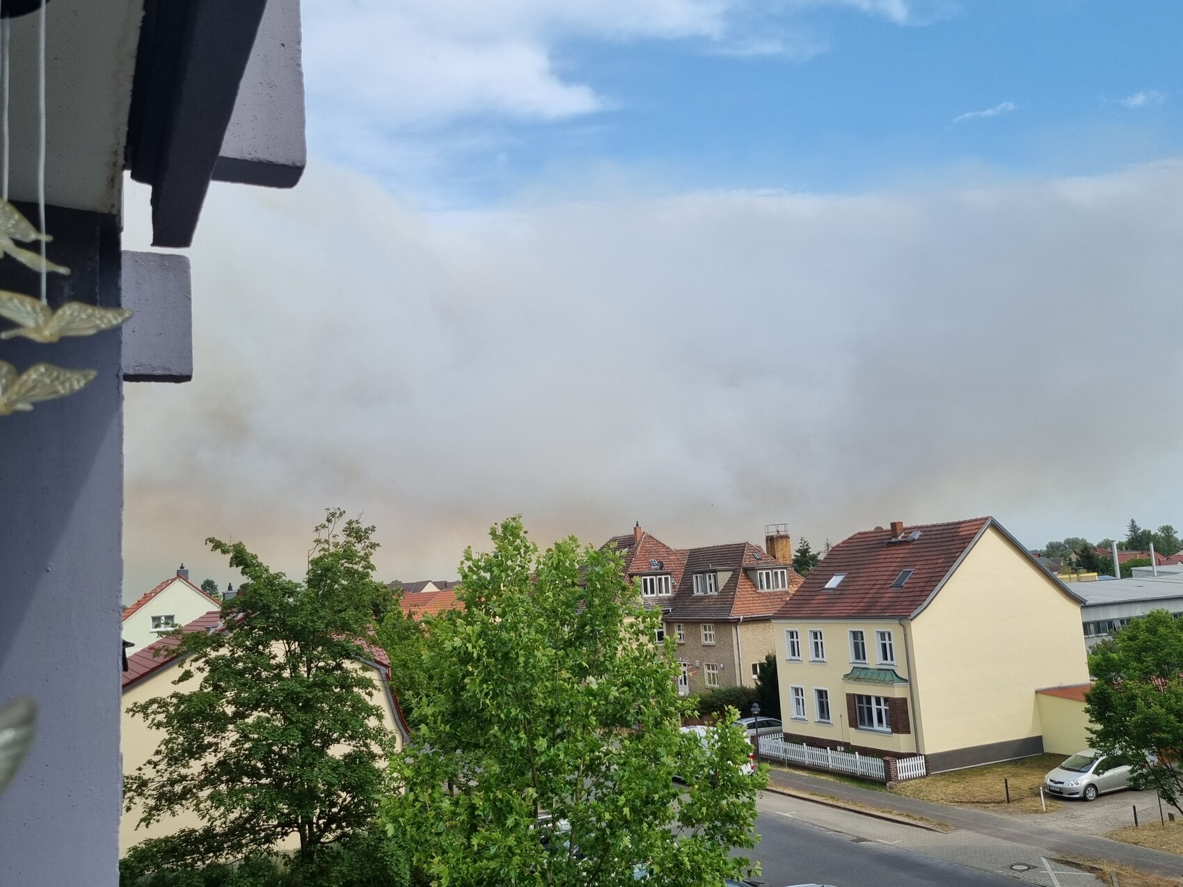 Wohnviertel mit Bäumen und roten Dächern; Rauch hinter den Häuserreihen am Horizont.
