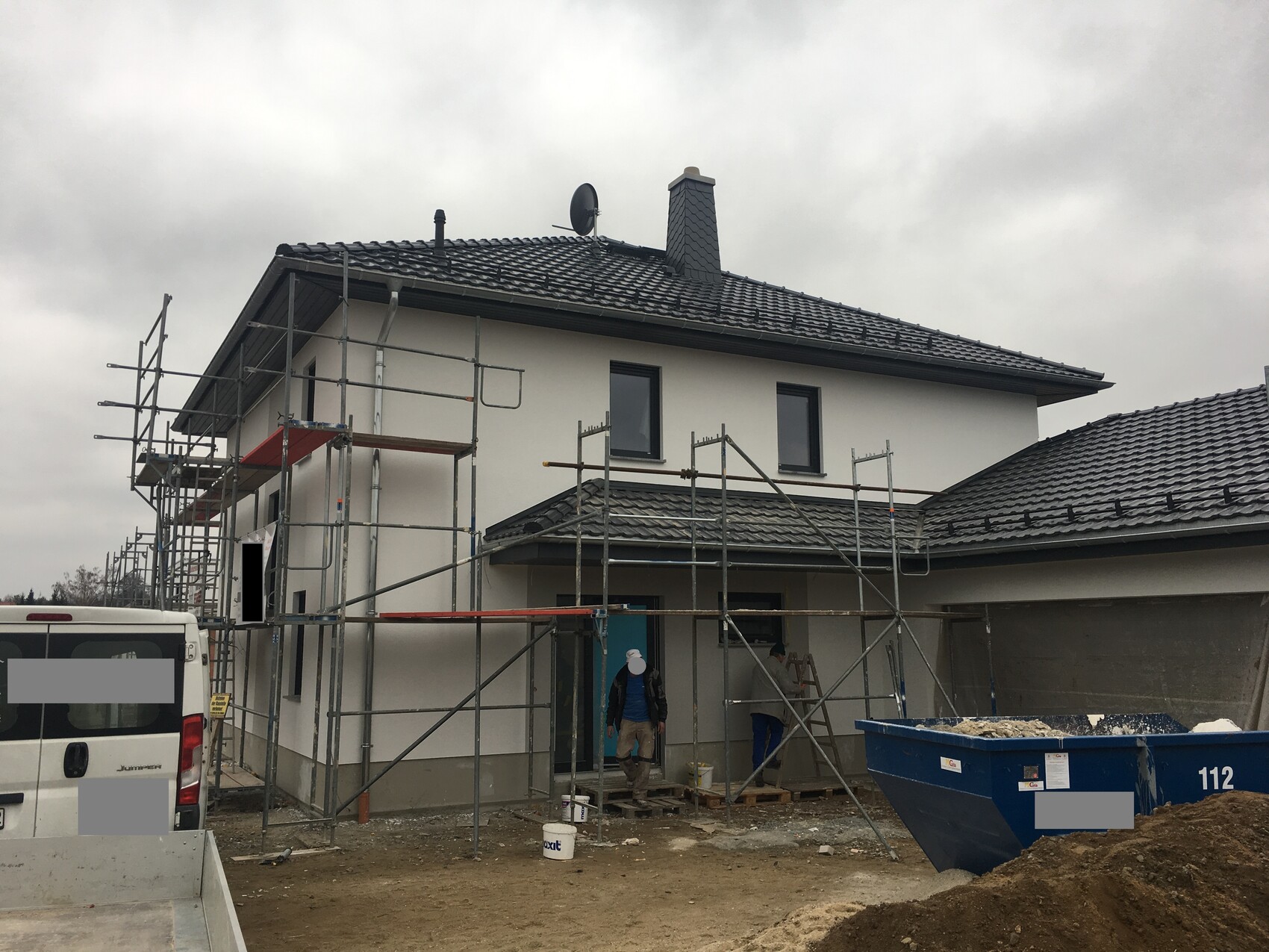 Zweistöckiges Haus im Bau mit Gerüst, Bauarbeitern und Bauschutt-Container.