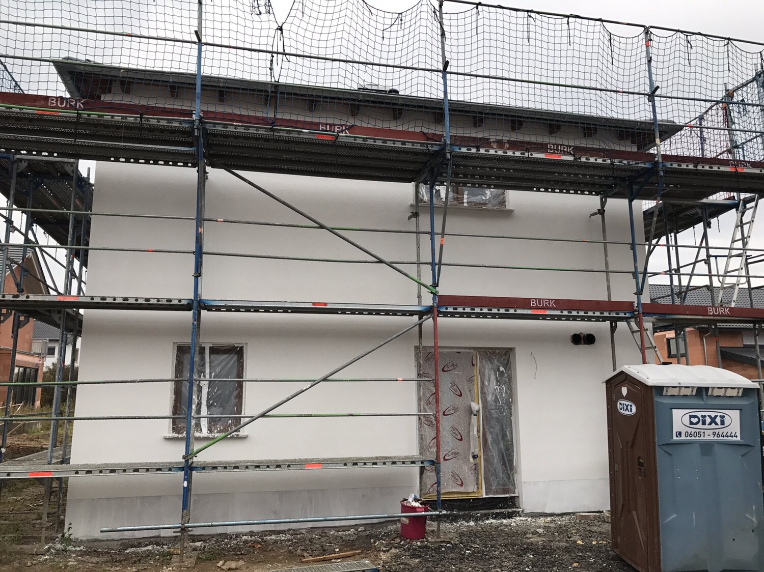 Weißes Haus im Bau, Gerüst mit Netzen, rechts Dixiklo.