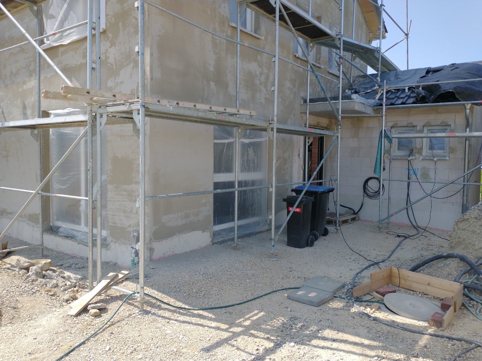 Baustelle: Haus mit Gerüst, Abdeckfolie an Fenstern, Müllcontainer und Schläuche.