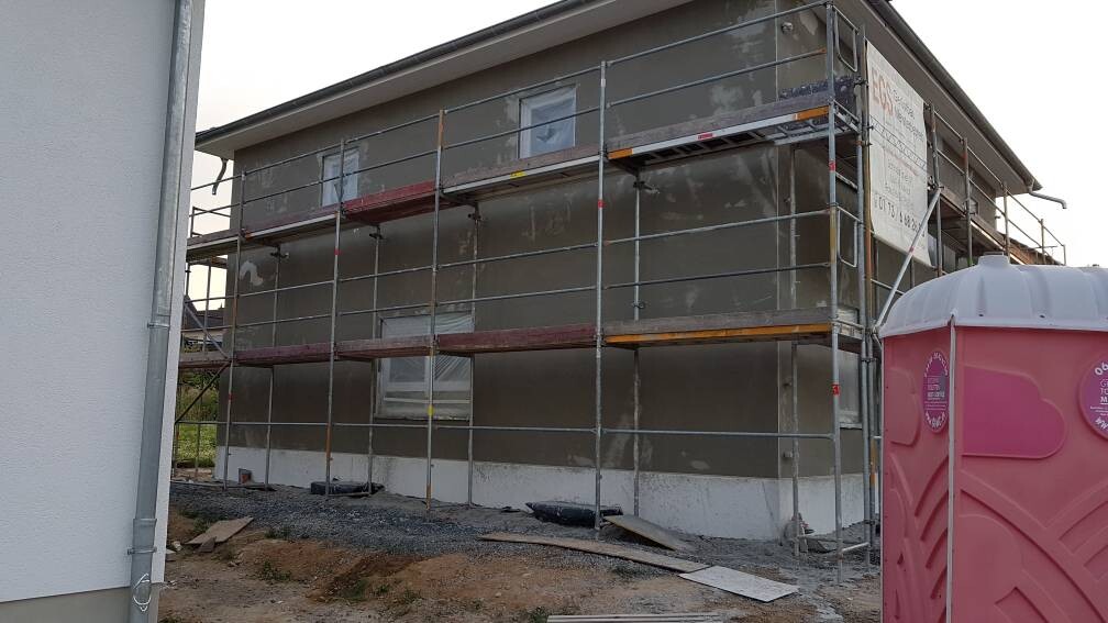 Außenansicht eines Hauses im Bau mit Gerüsten; rechts eine pinke Baustellentoilette.