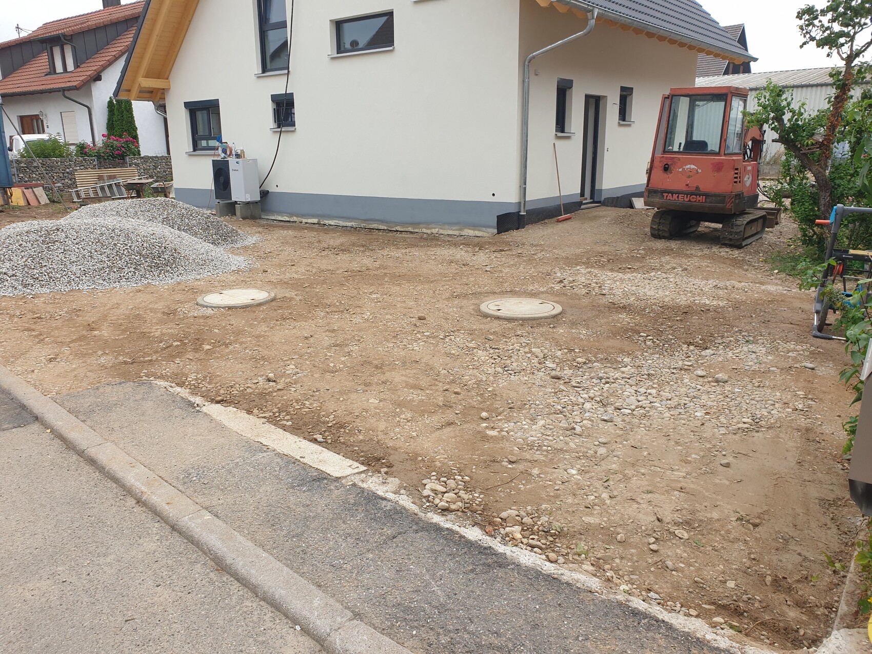 Baustelle vor Wohnhaus: unbefestigter Hof, Kieshaufen, Takeuchi-Bagger, zwei runde Kanaldeckel.