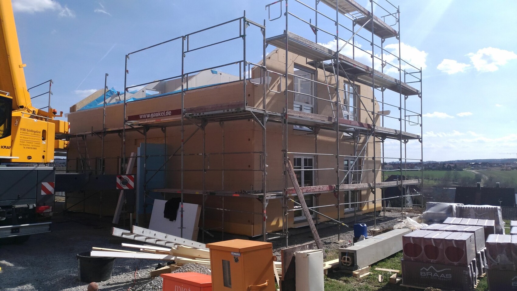 Bau eines zweigeschossigen Holzhauses mit Gerüst und Baustellenmaterialien; Kran links sichtbar.