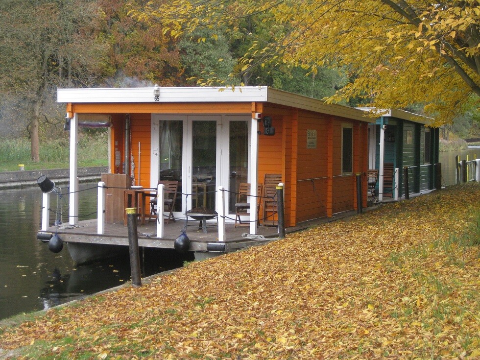 Buntes Hausboot mit Terrasse am Wasser, Herbstlaub am Uferweg.
