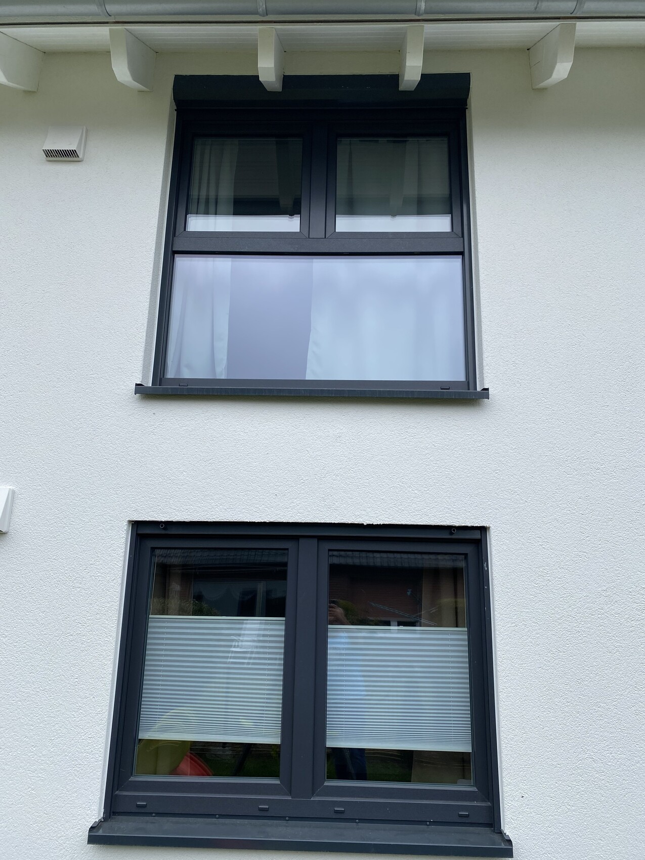 Zwei dunkel gerahmte Fenster übereinander an einer weißen Fassade; obere Vorhänge, unten Jalousien.