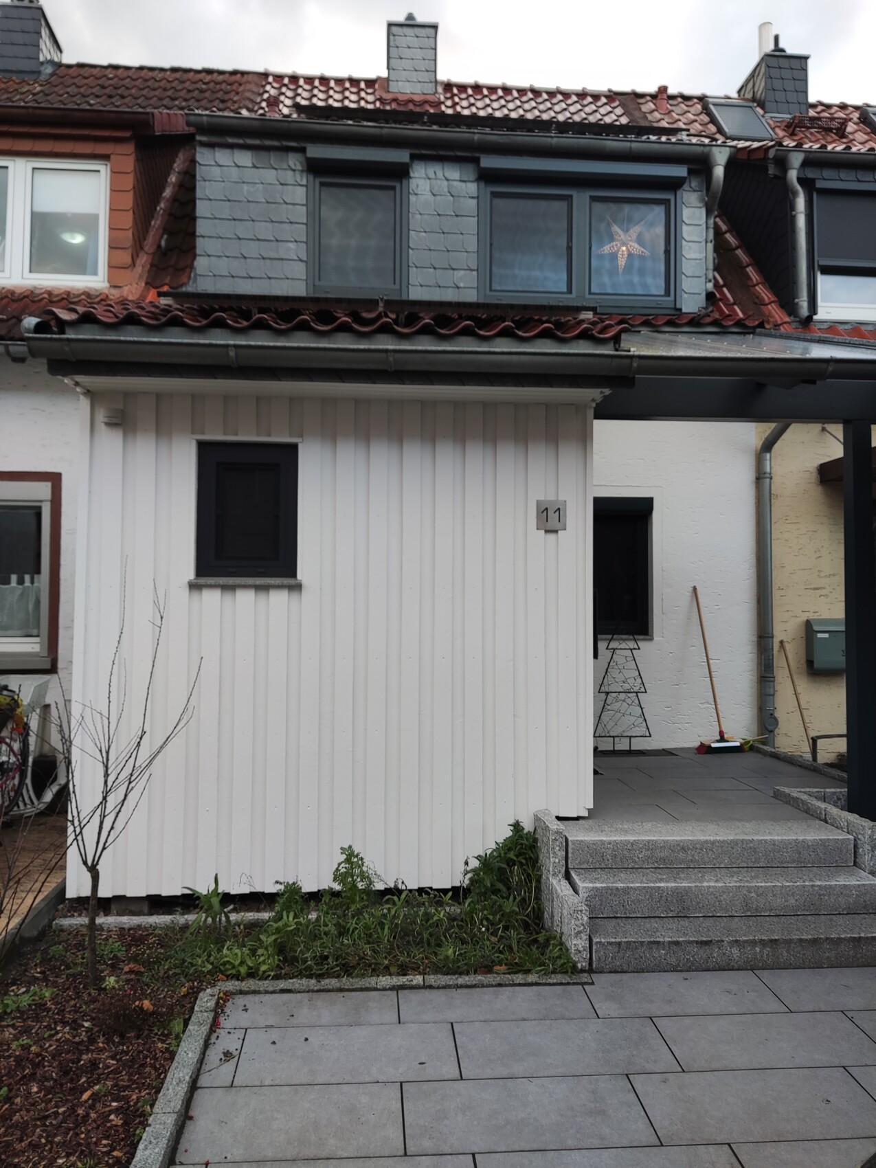 Außenfront eines Hauses: weiße Wandverkleidung, Treppe, Pflanzenbeet, Hausnummer 11.