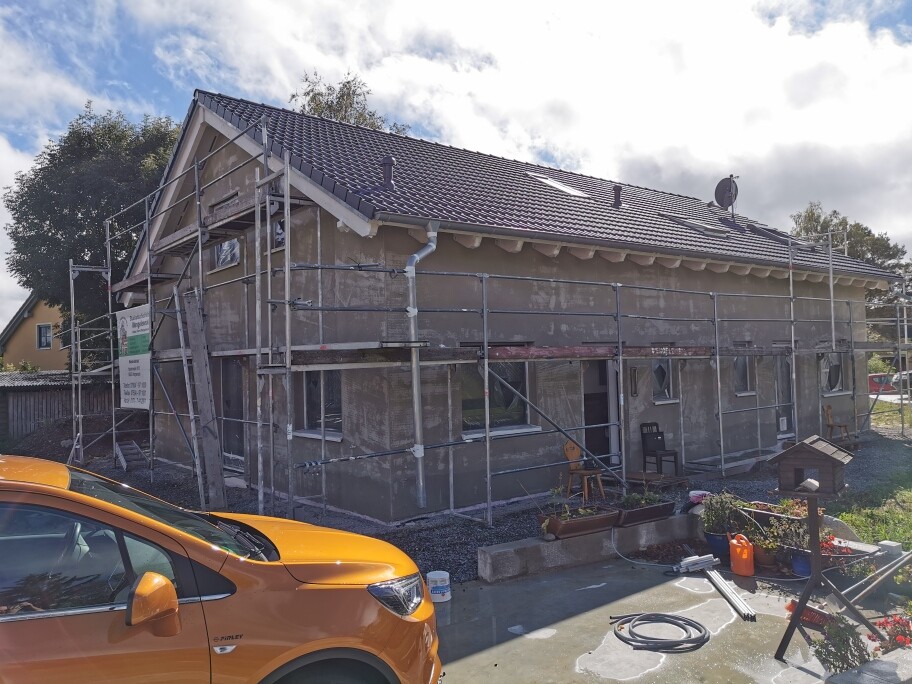 Ein Einfamilienhaus wird saniert, umlaufendes Gerüst um das Gebäude; orangefarbener Pkw im Vordergrund
