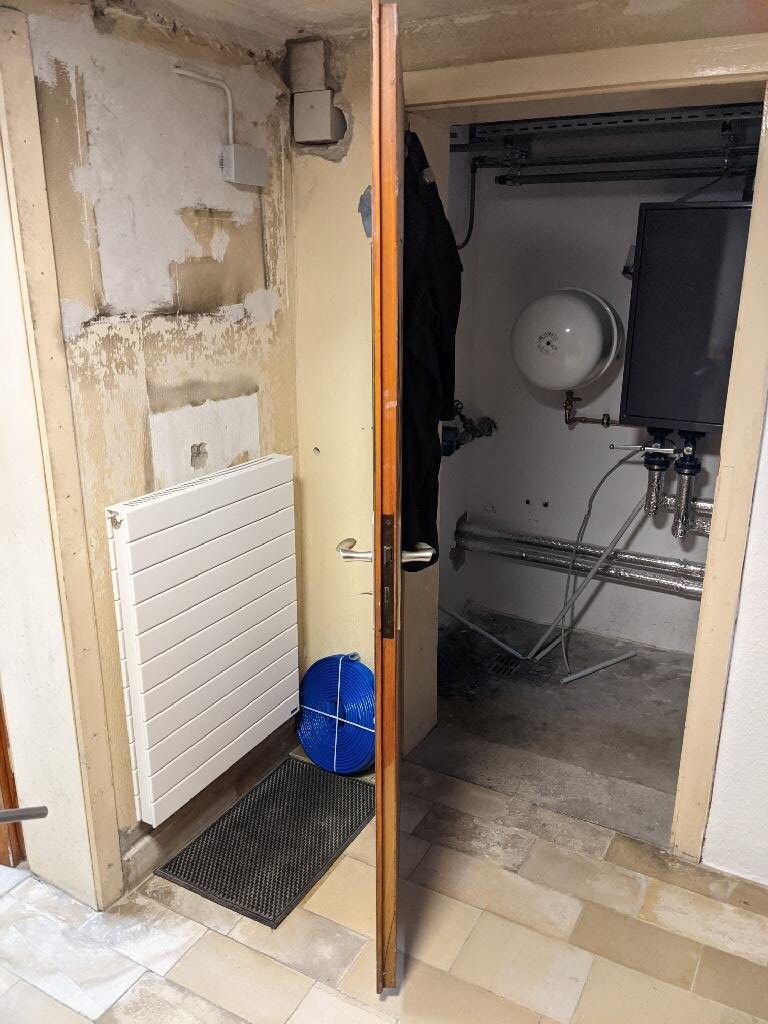 Offener Heizungsraum mit Boiler und Expansionsgefäß, sichtbare Rohre und blaue Schlauchrolle.
