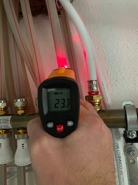 Hand hält Infrarot-Temperaturmesser vor Rohrleitung mit Ventilen; Display 23,2°C, roter Laser.