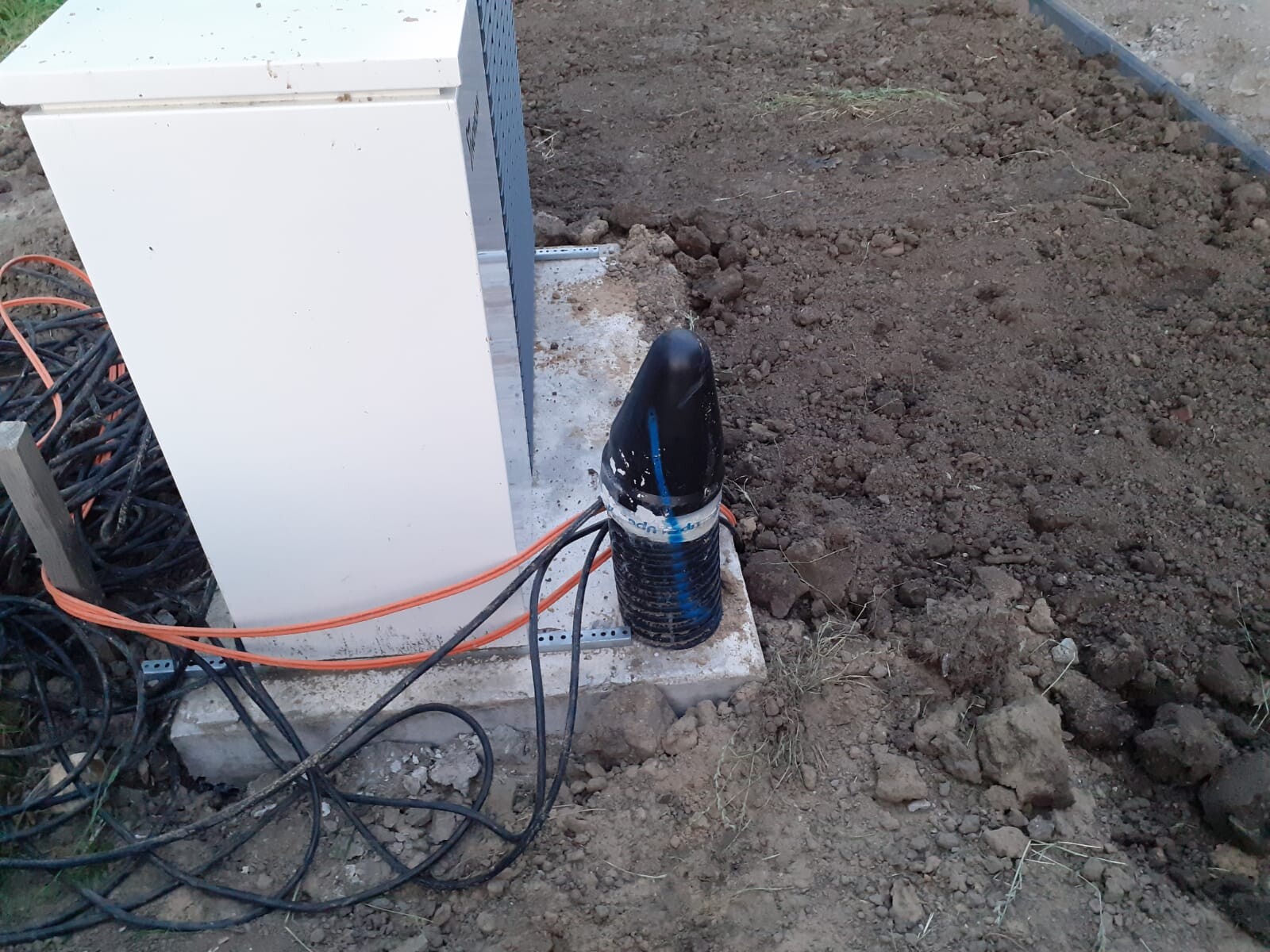 Außen-Klimagerät auf Betonfundament, Kabelsalat aus schwarzen und orange Kabeln im Erdreich.