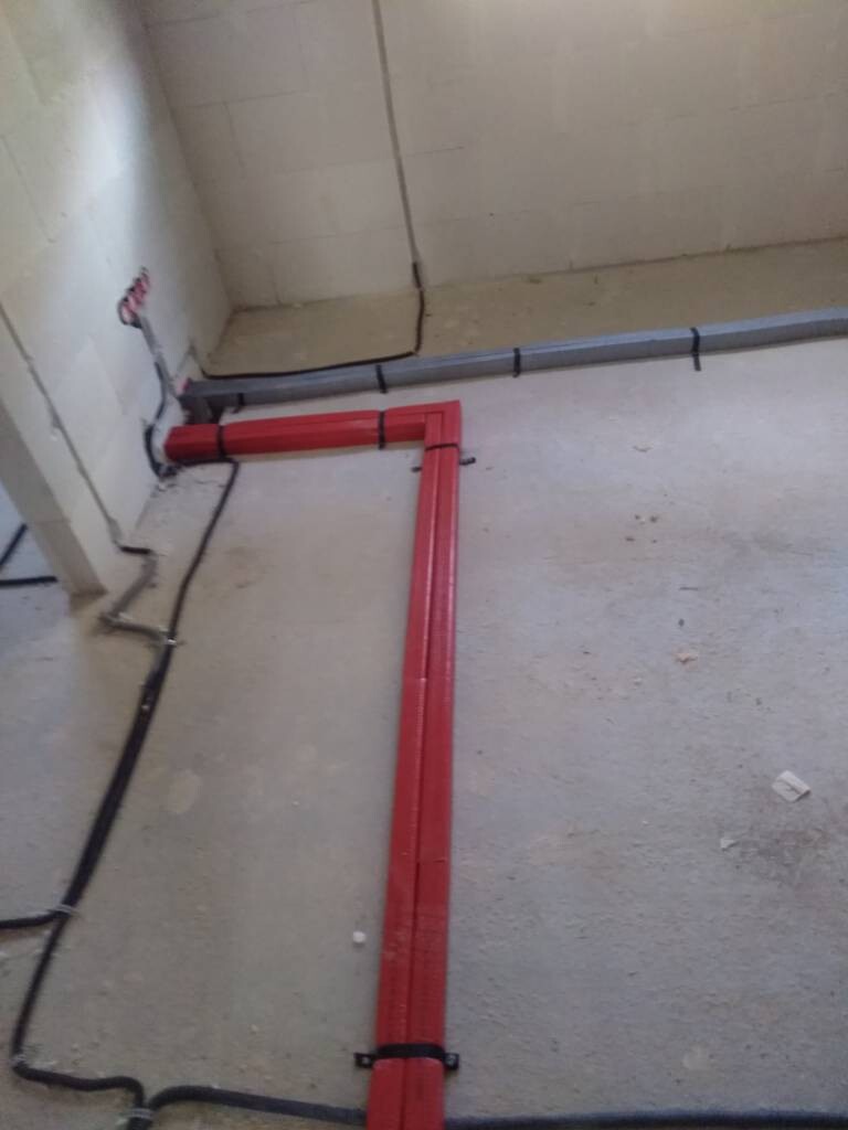 Rote Kunststoffrohre bilden eine L-förmige Leitung auf dem Betonboden, daneben schwarze Kabel.