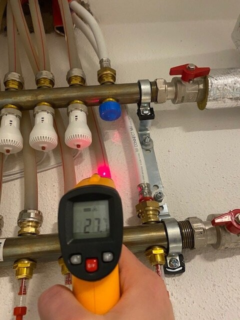 Hand hält Infrarot-Thermometer vor Heizungsrohren mit Ventilen; Anzeige 27°C.