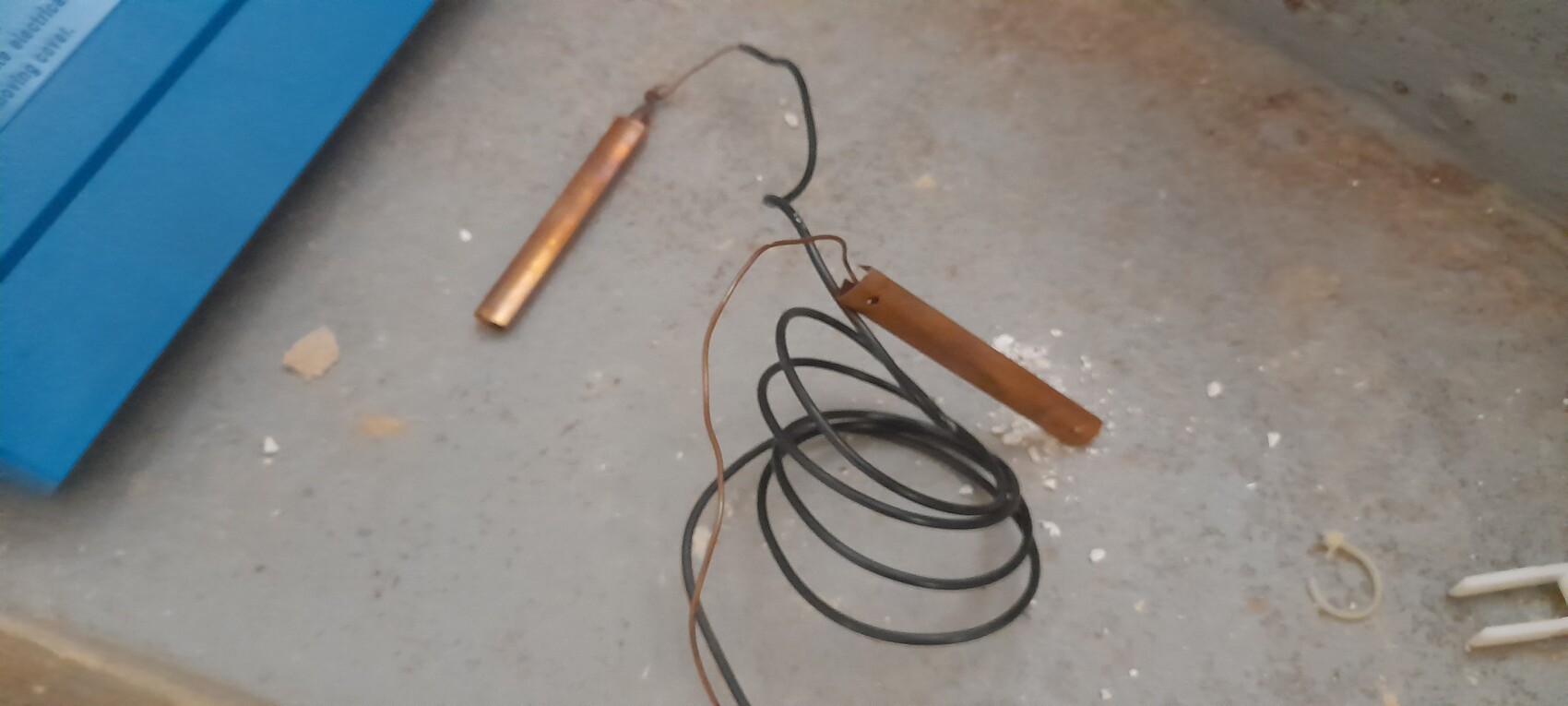 Auf Betonboden liegt schwarzes Kabel, daneben Kupferdraht und rostbraune Stange.