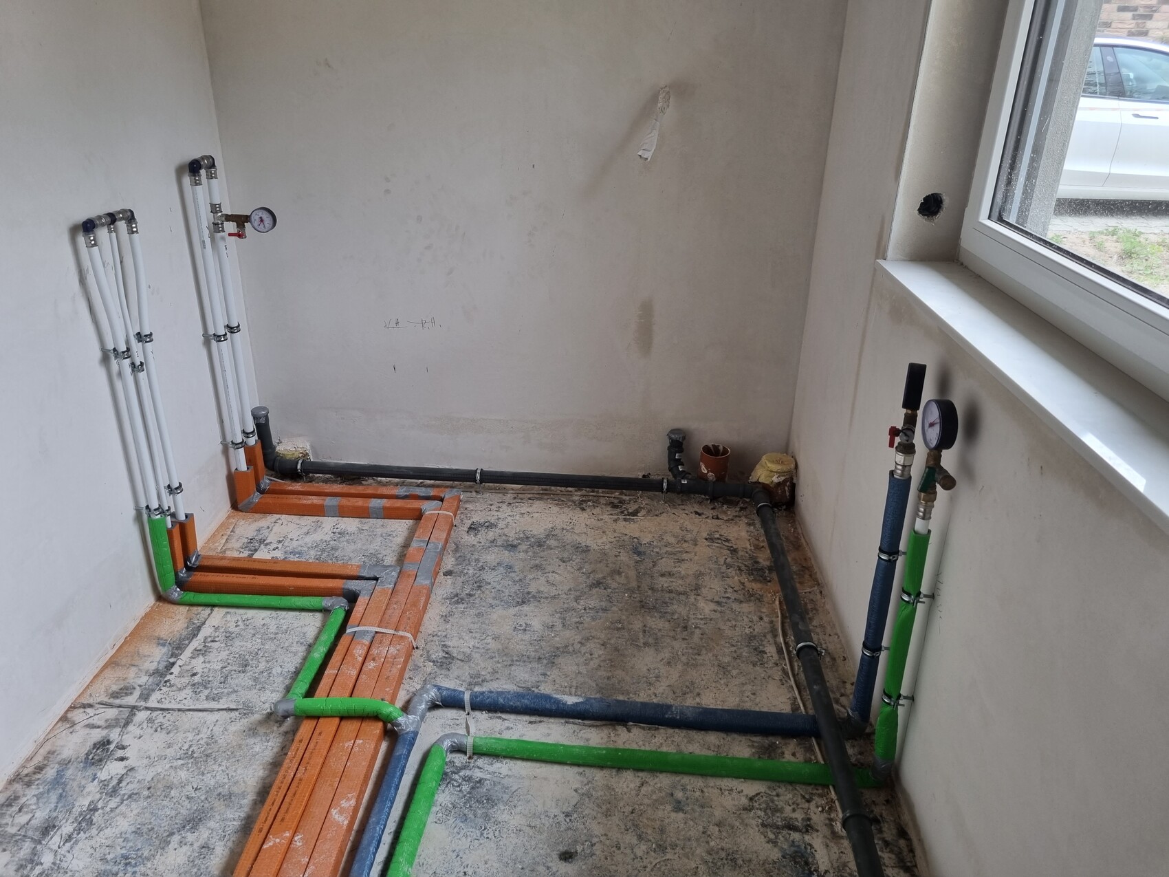 Installationsraum mit farbigen Wasserrohren (orange, grün, blau) am Boden, Fenster rechts.