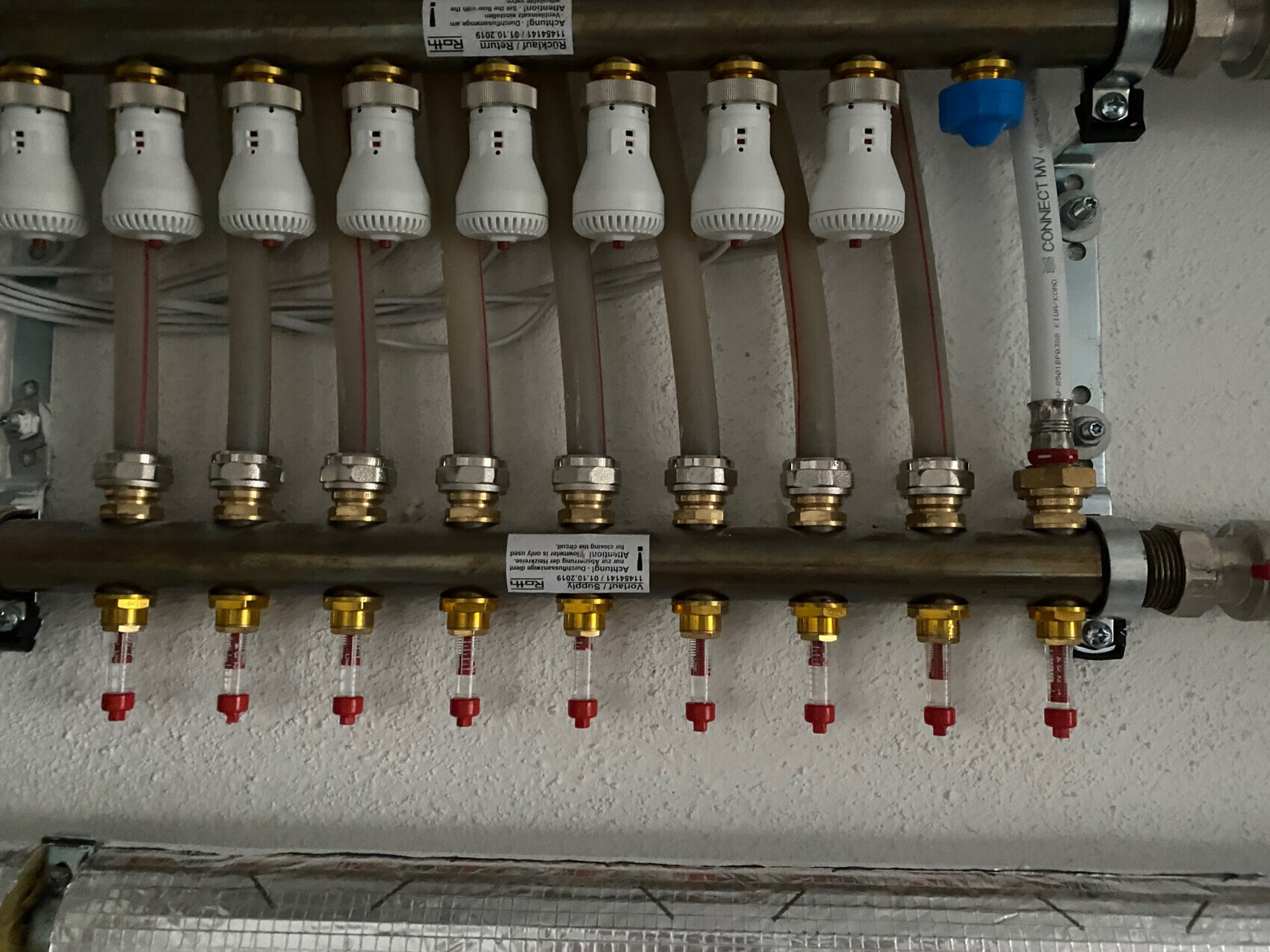 Heizungs-Verteiler mit mehreren Leitungen, weißen Thermostatventilen und Messingkupplungen.