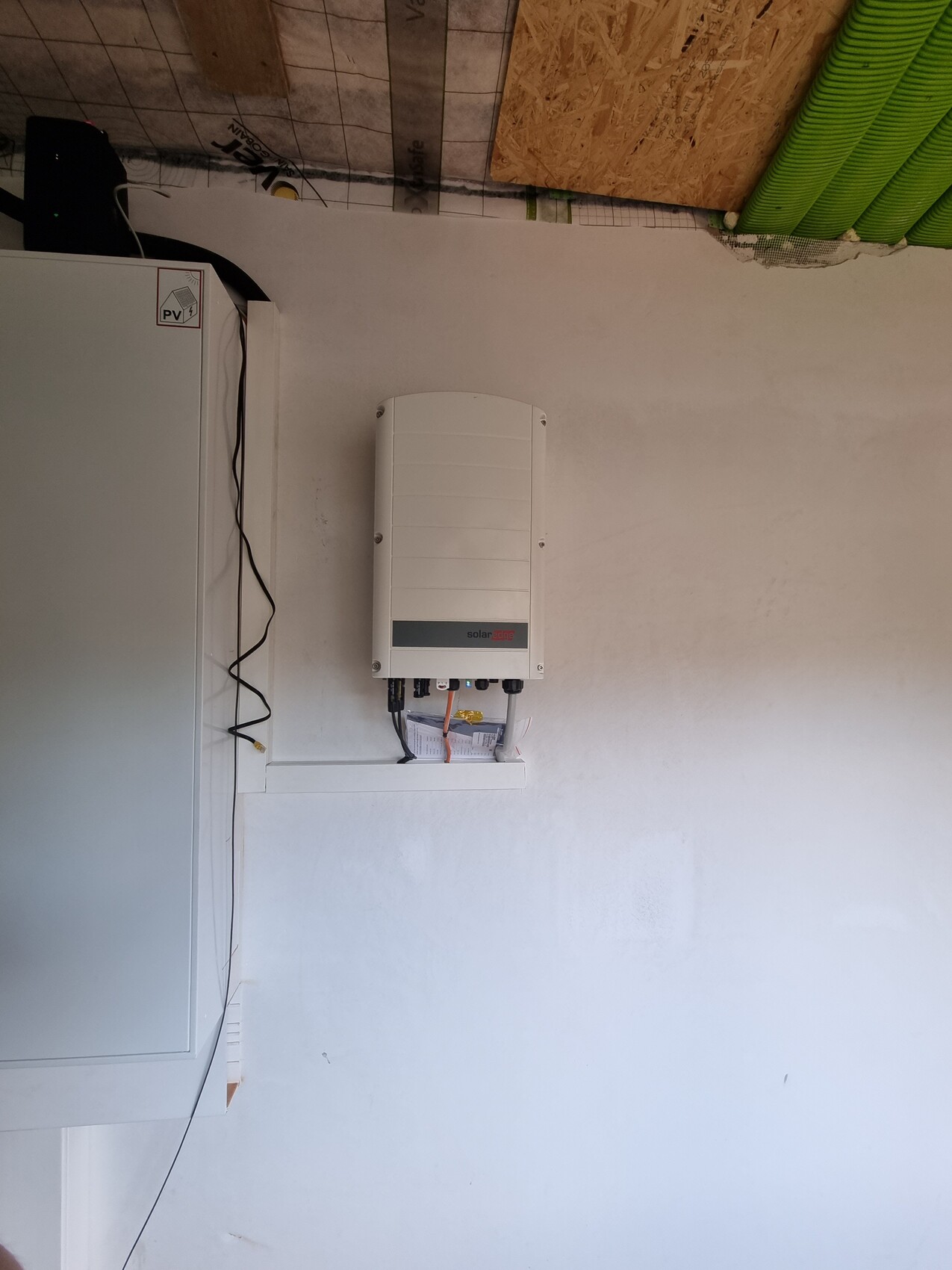 Weißer Solar-Wechselrichter an der Wand, Kabelstränge laufen nach unten neben Schaltschrank.