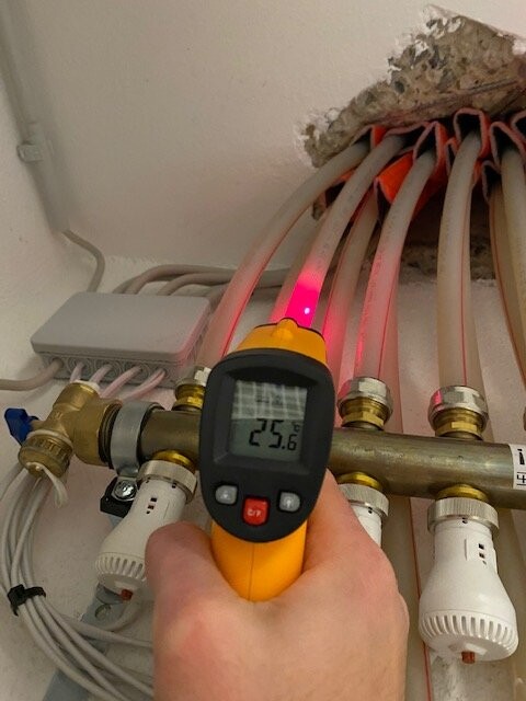 Hand hält digitales Messgerät mit Laser auf Heizungsrohr; Anzeige 25,6°C.