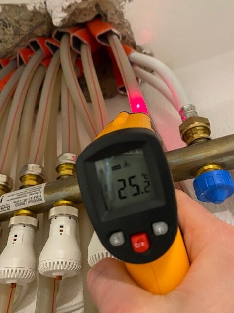 Hand hält Infrarot-Thermometer an Heizungsrohre; Display zeigt 25,2°C.