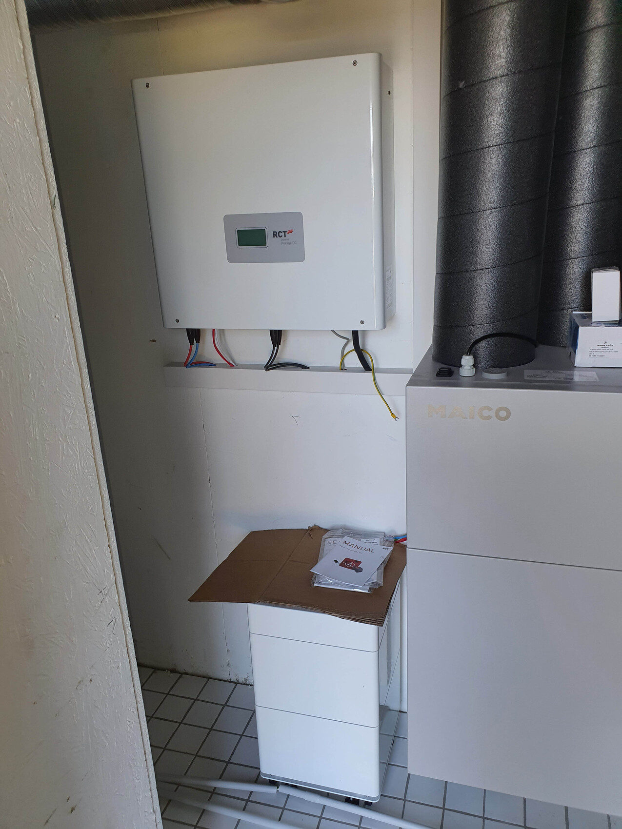MAICO Lüftungsgerät rechts, Wandgerät mit Kabeln links, Karton mit Anleitung auf Schrank.